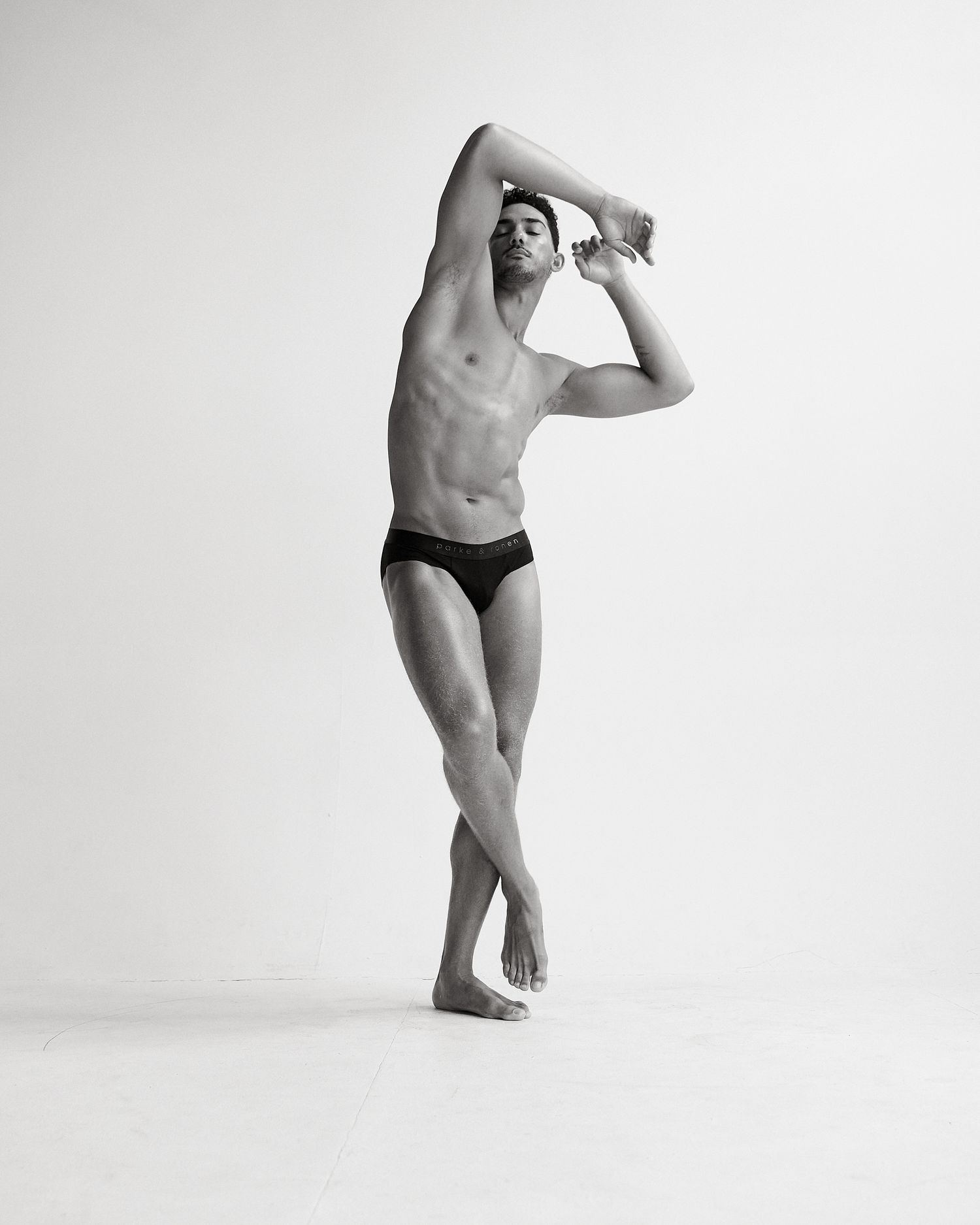 Black and white photo of a muscular man in swim briefs posing gracefully with arms raised overhead.
