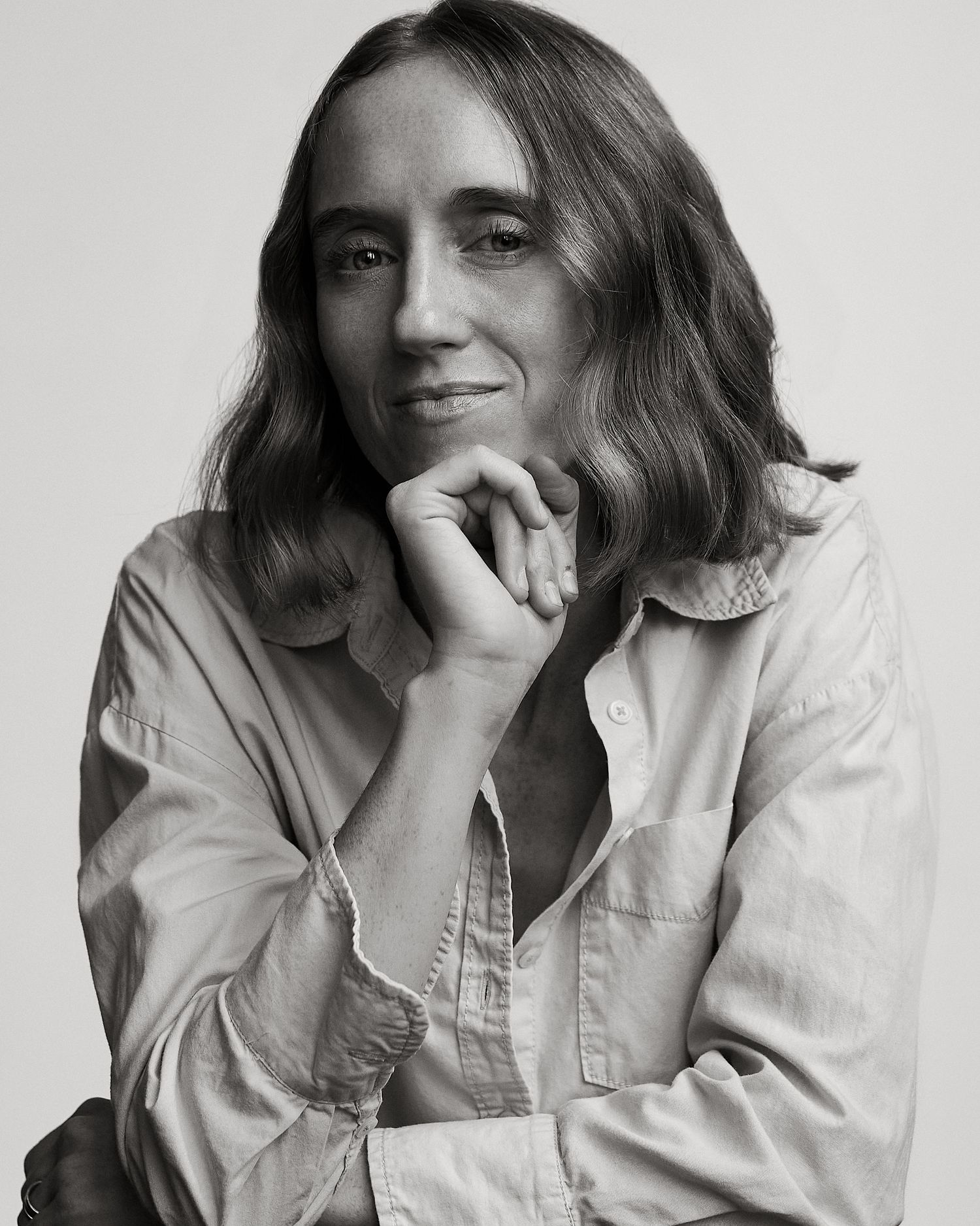 Black and white portrait of a person with shoulder-length wavy hair, wearing a light button-up shirt, hand on chin.