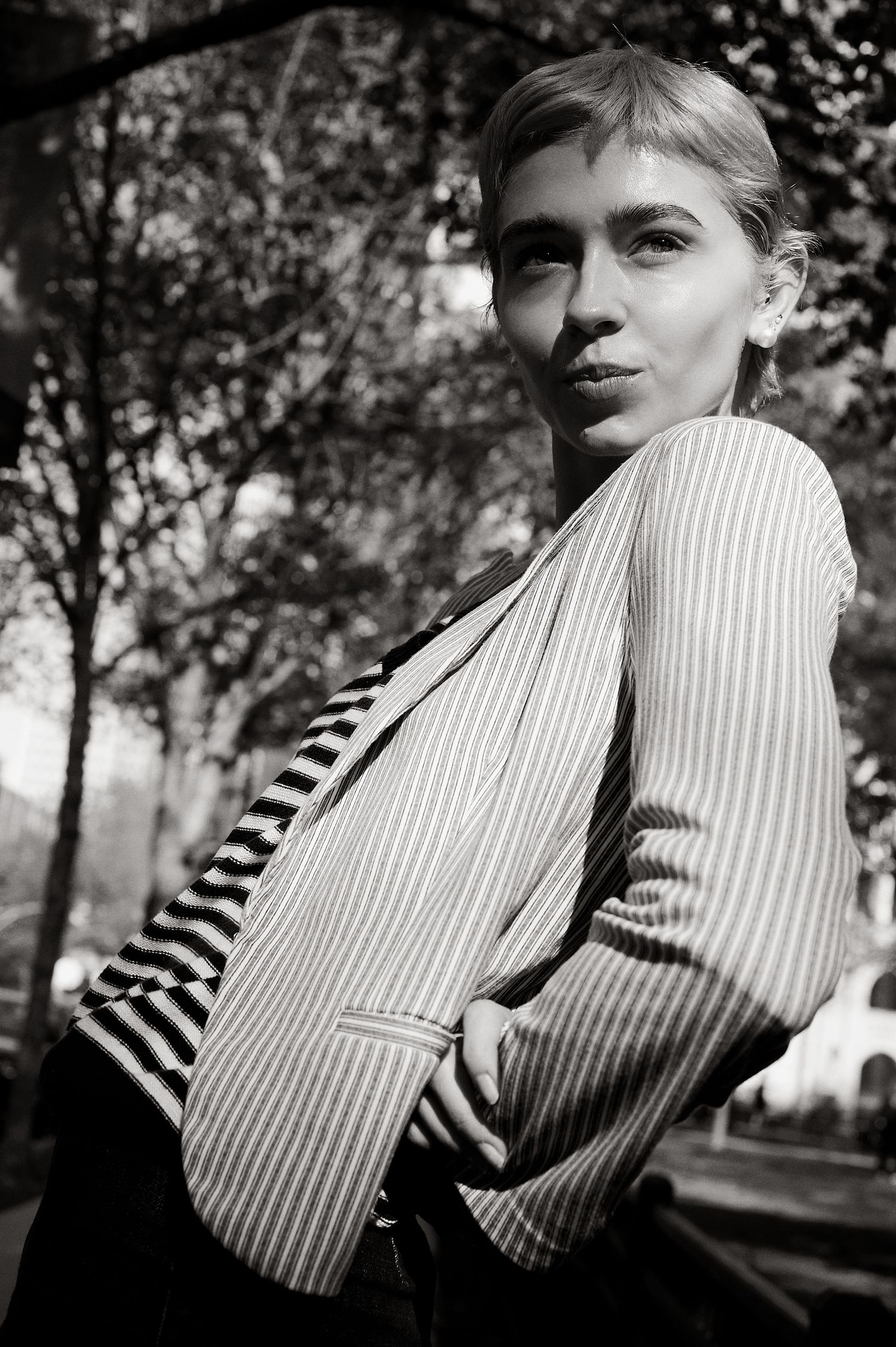 Young blonde woman in pinstripe blazer poses confidently among trees in moody black and white outdoor portrait.