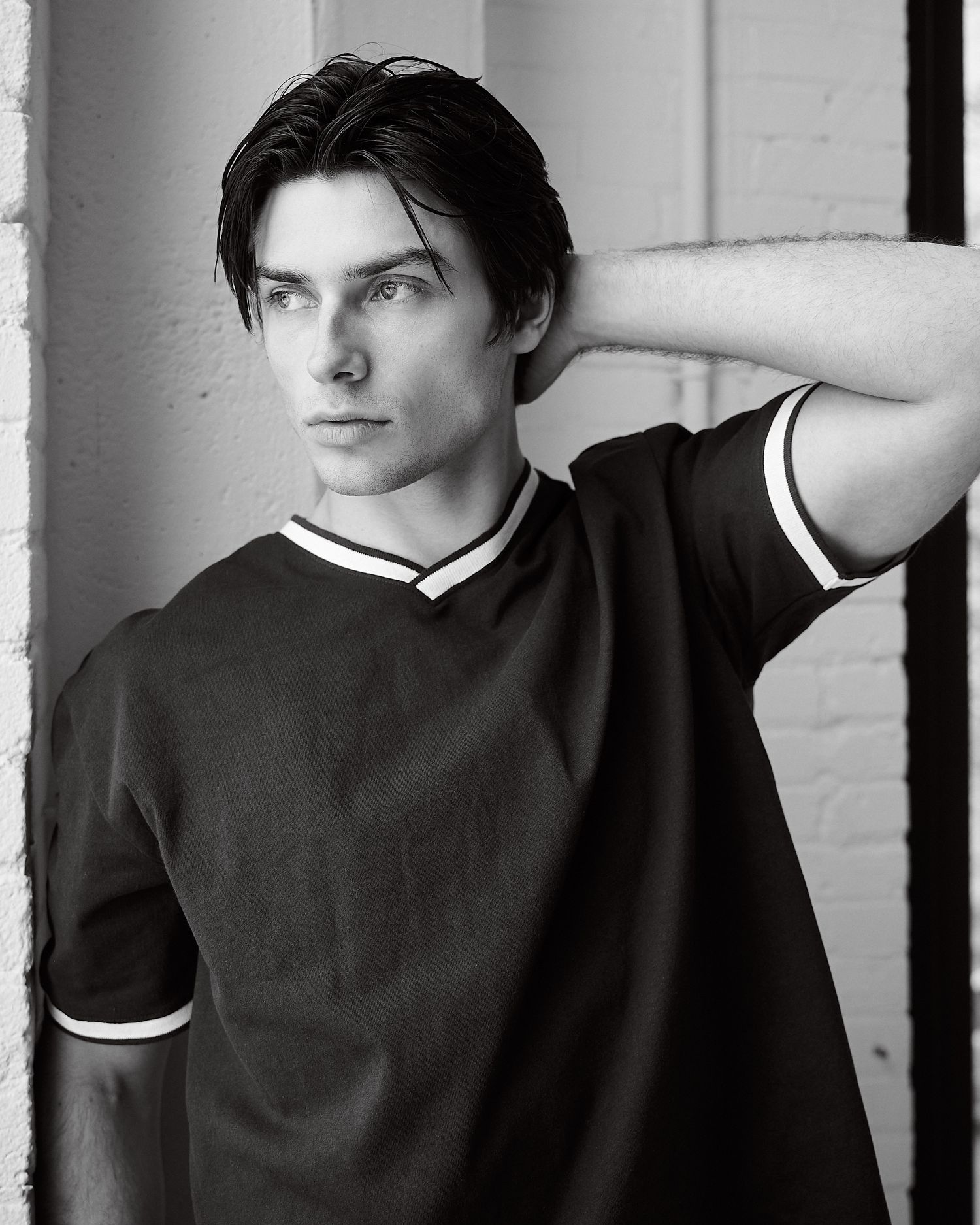 Black and white portrait of a young man in a dark V-neck t-shirt posing with hand behind his head.
