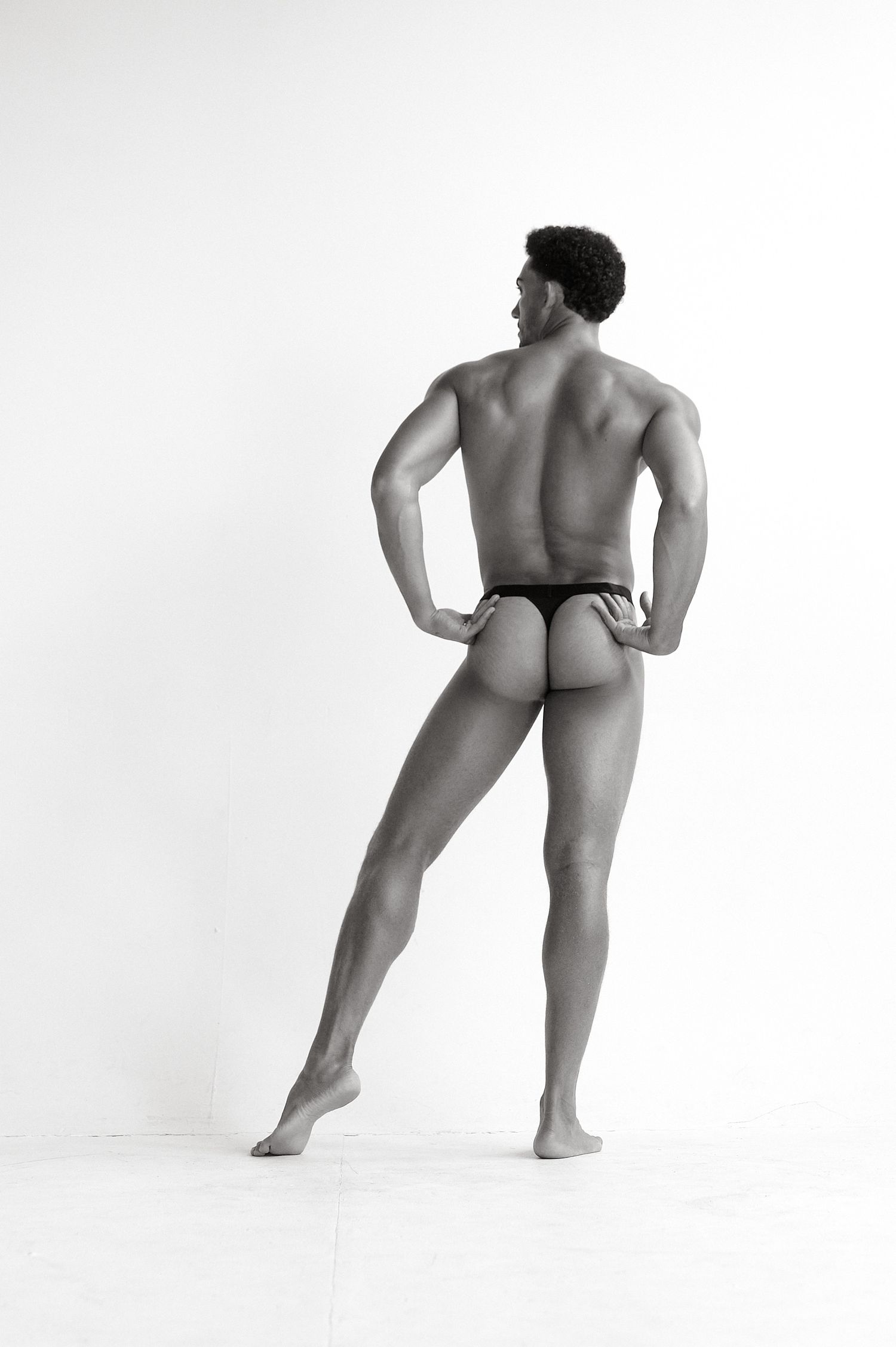 Rear view of athletic male model in black thong standing with hands on hips in a white studio.