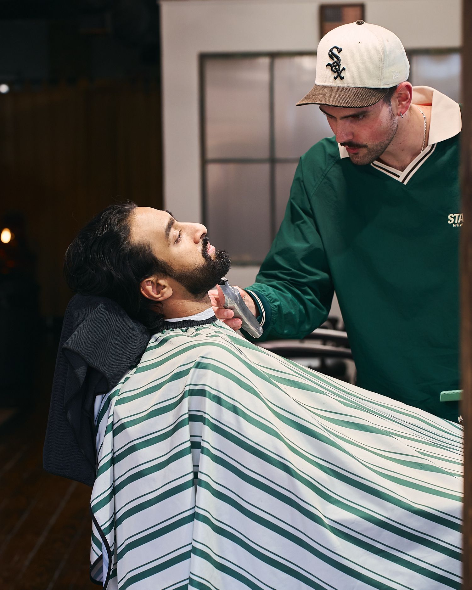 Skilled barber in White Sox cap trimming a bearded clients facial hair with a razor.