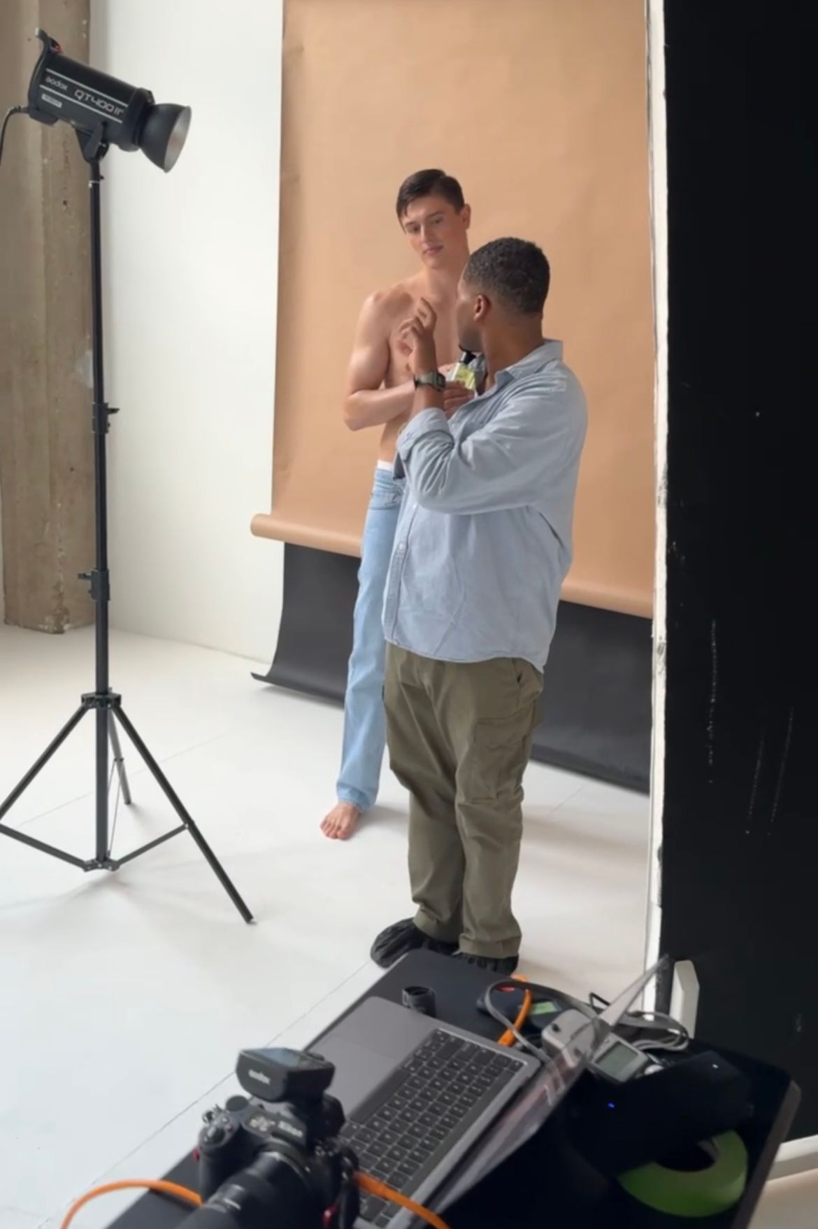 Behind-the-scenes photo shoot with a shirtless male model being styled in a professional studio.