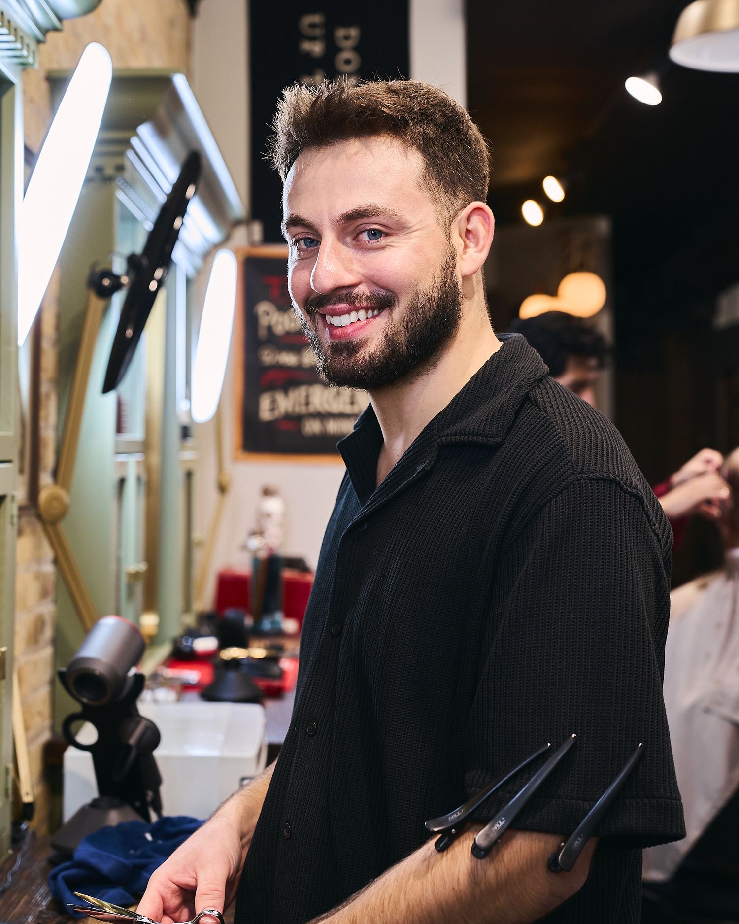 Smiling bearded barber in black shirt holding scissors inside a stylish barbershop.