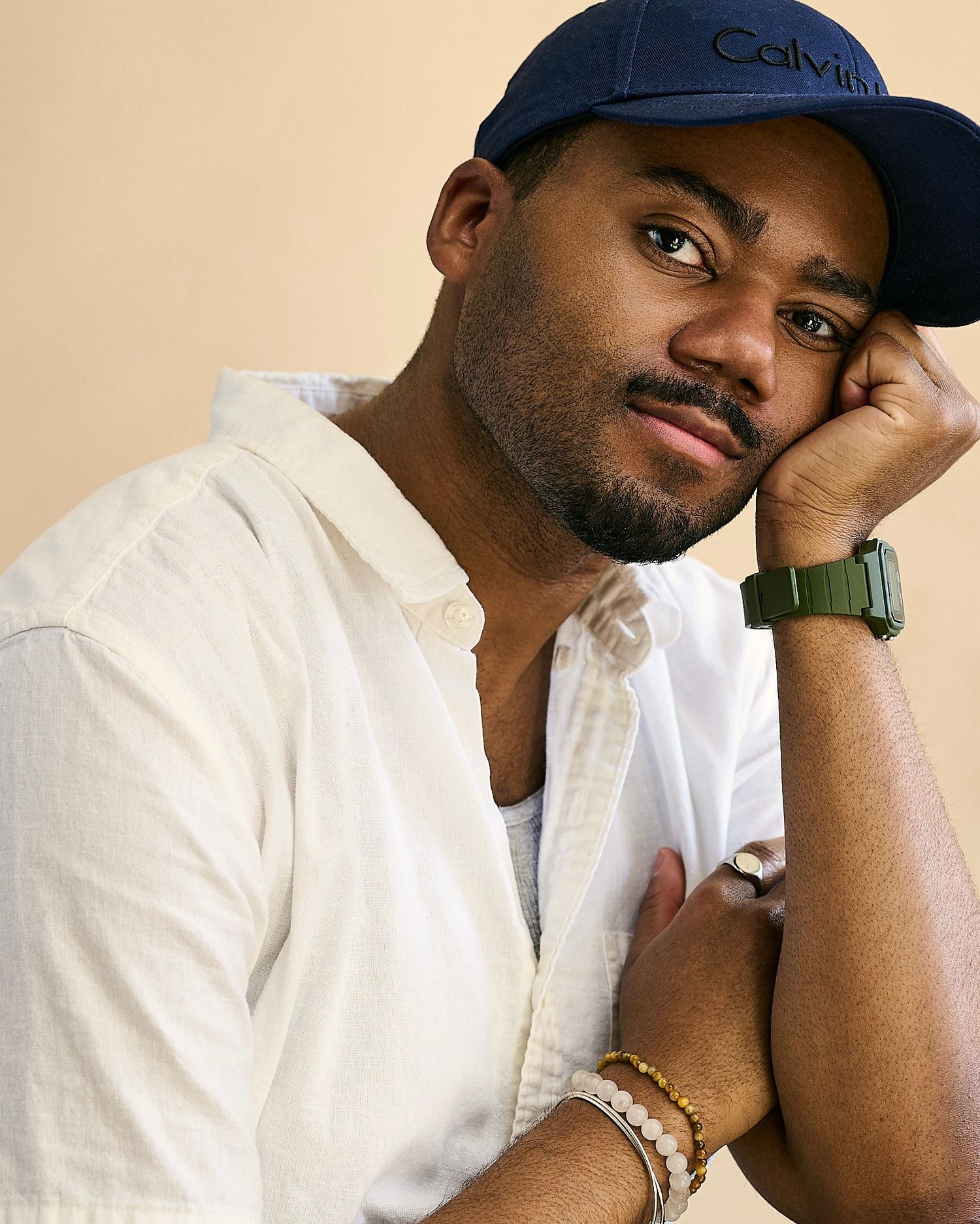 Man in Calvin Klein cap and white shirt poses thoughtfully, wearing a green watch and beaded bracelets.