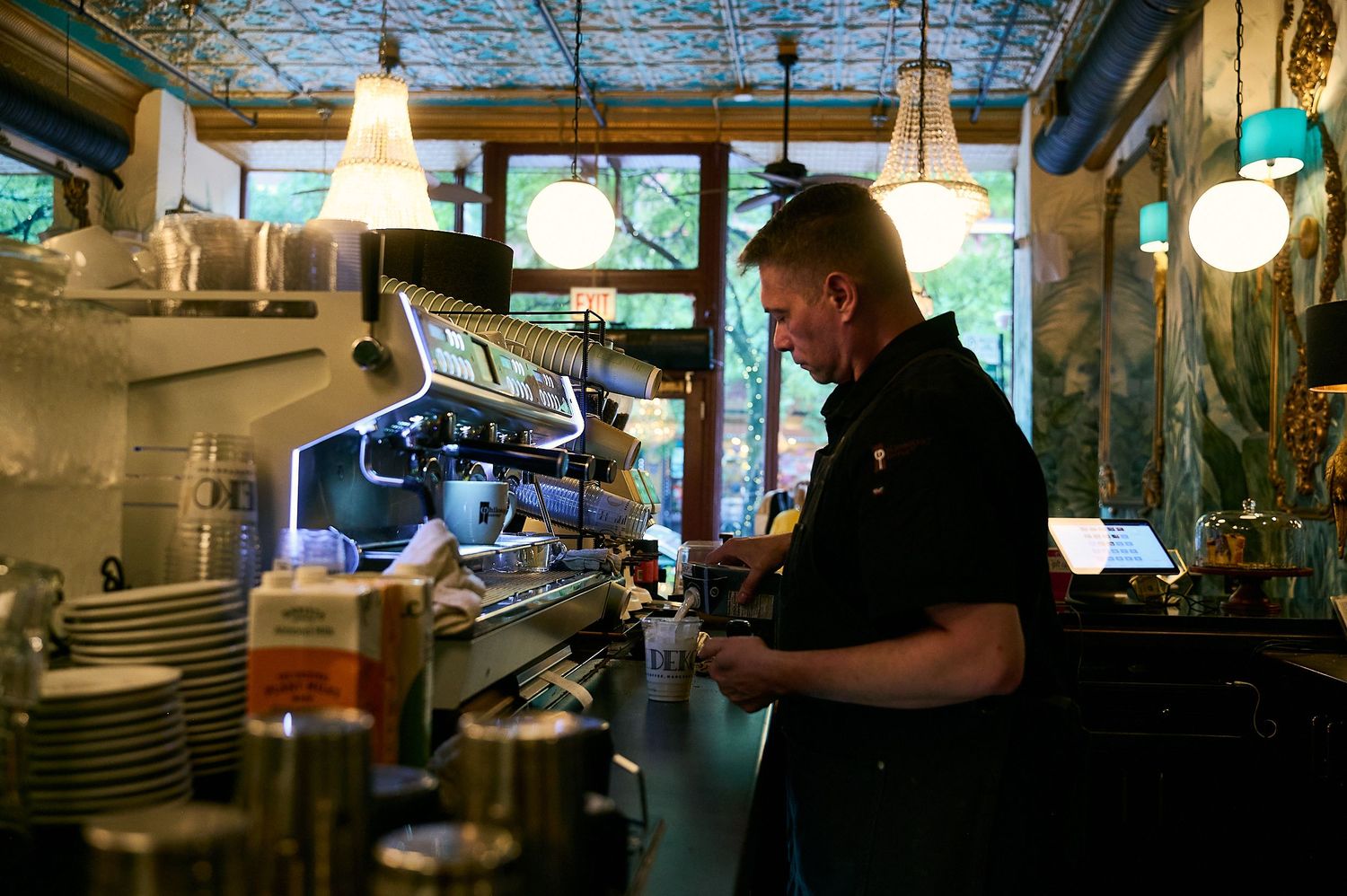 Barista working behind an espresso machine in a cozy cafe with vintage decor and pendant lights.