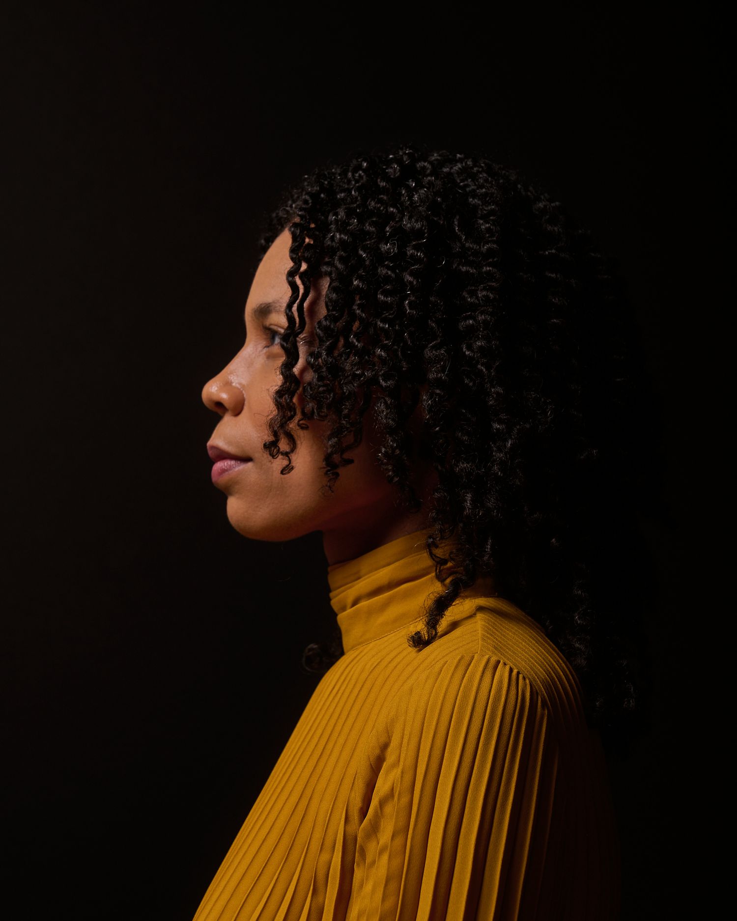 Side profile of woman with curly hair wearing yellow turtleneck against dark background.