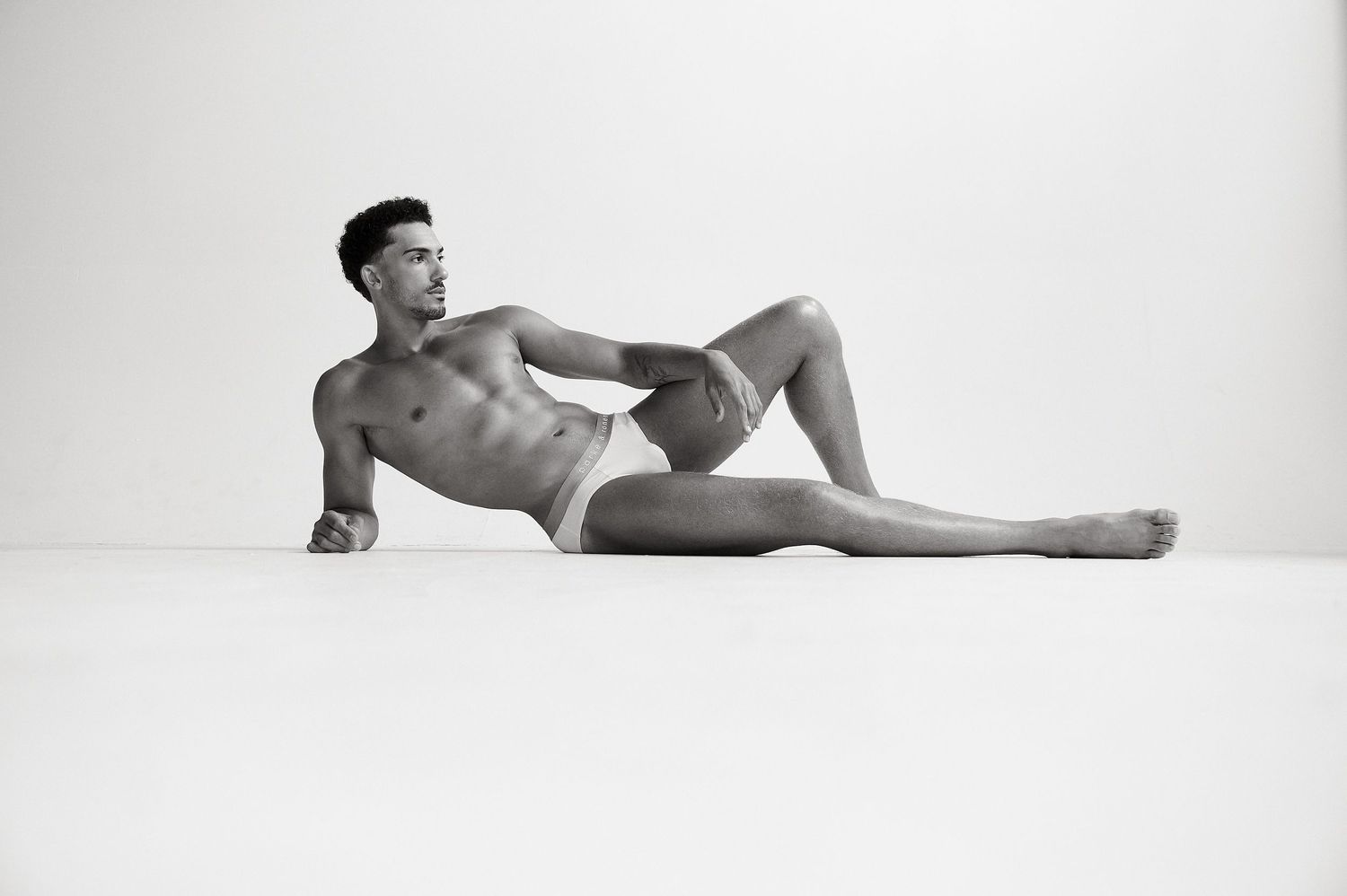 Athletic male model in briefs lying on a white studio floor in a relaxed pose, black and white photography.
