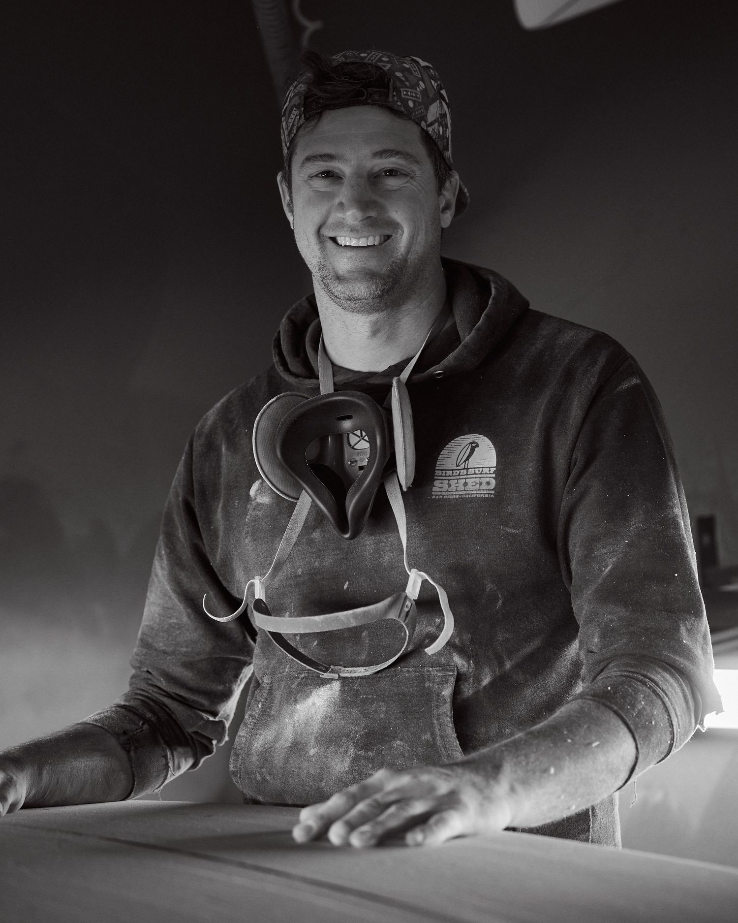 A smiling surfboard shaper covered in foam dust works in his shaping workshop.