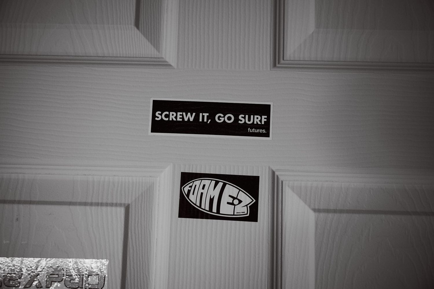 A door sign reading Screw It Go Surf above a Firewire Surfboards oval logo sticker.