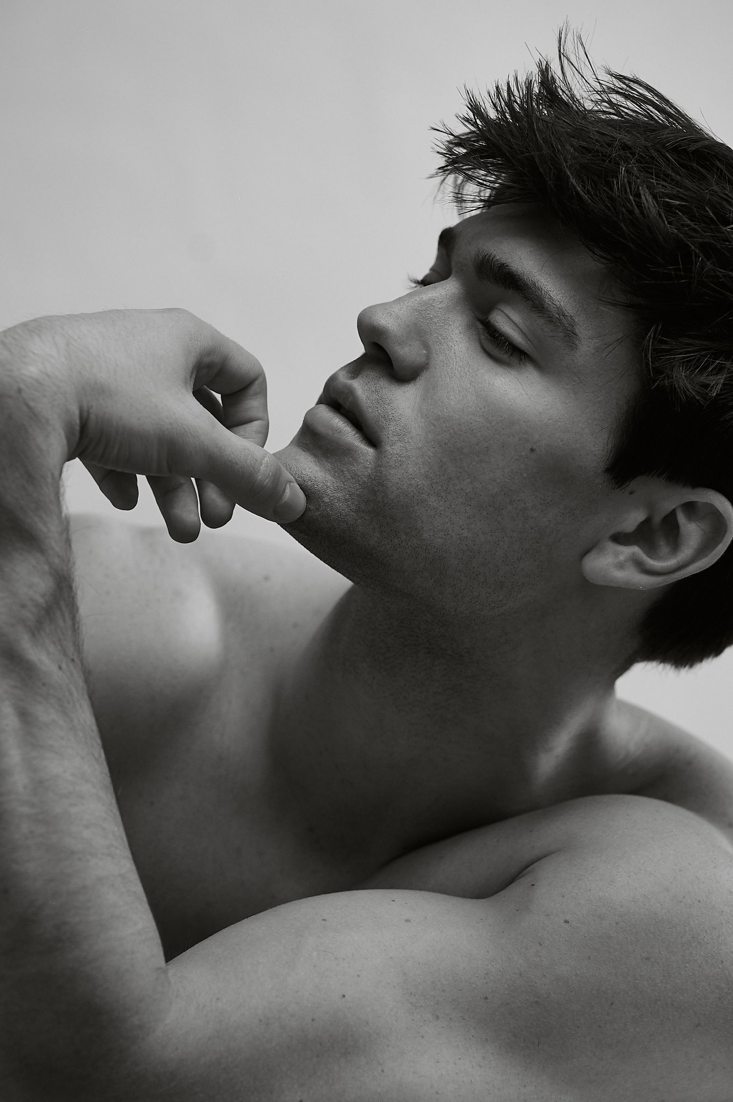 Black and white portrait of a shirtless muscular man with dark hair touching his chin with eyes closed.