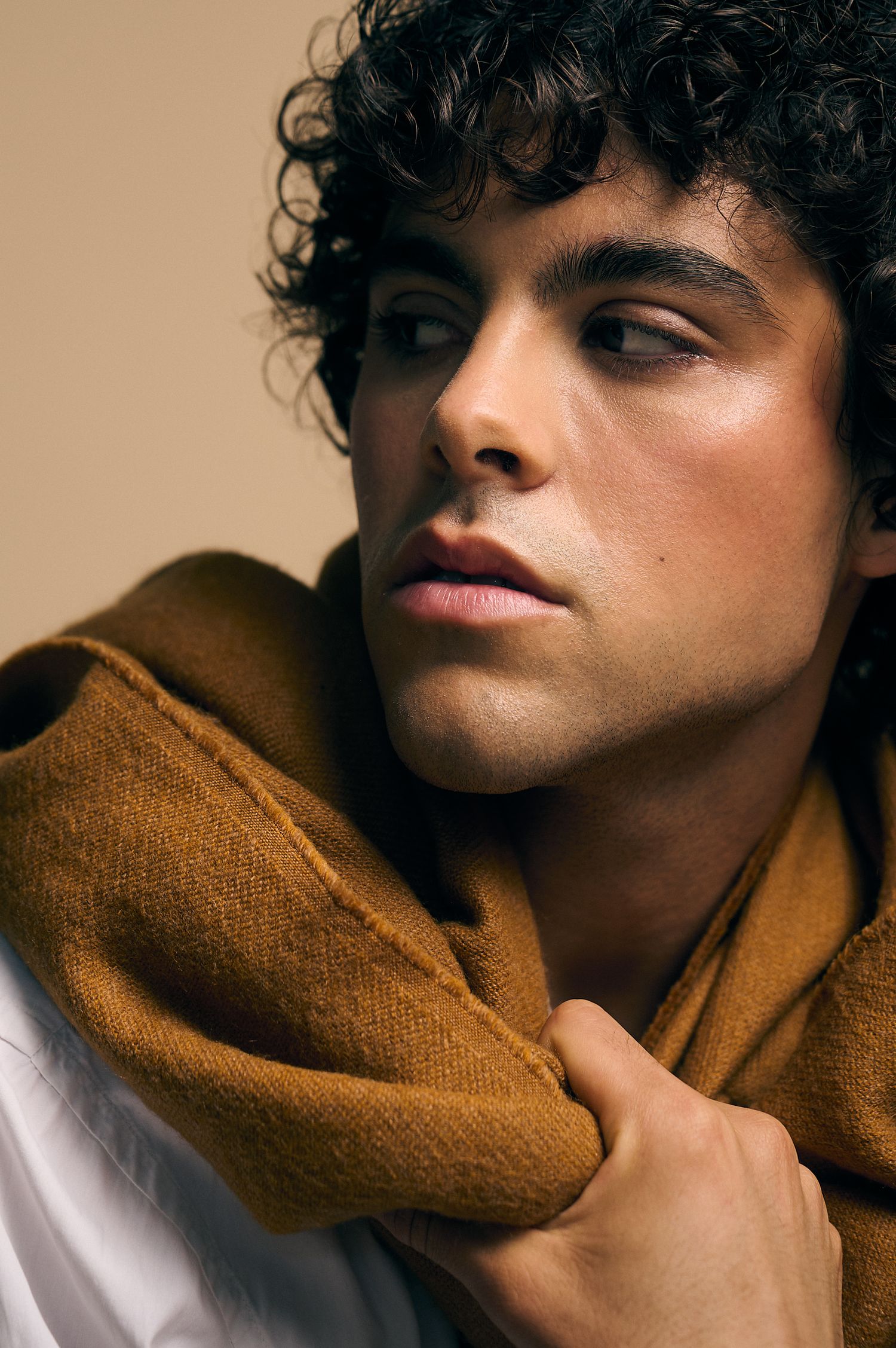 Young man with curly dark hair wearing a mustard yellow scarf and white shirt, gazing thoughtfully to the side.