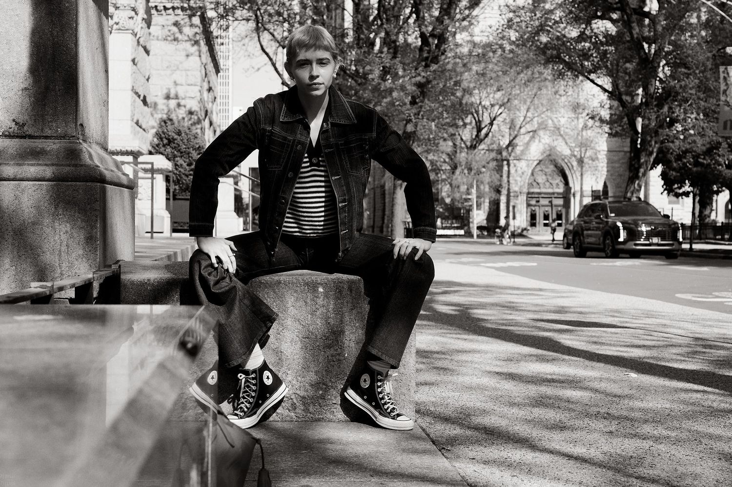 Young woman in denim jacket and Converse high-tops sits on stone fixture in urban park, black and white photo.