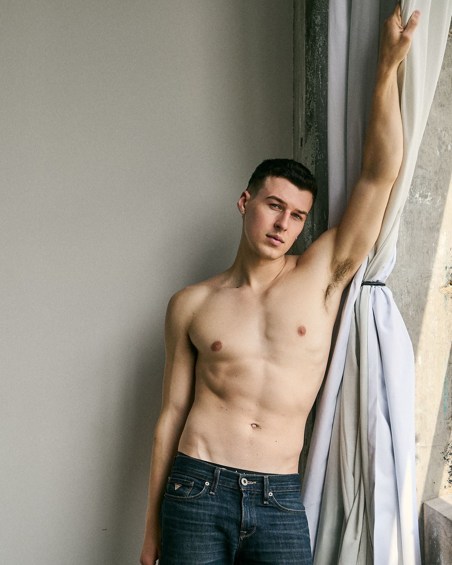 Shirtless young man in jeans posing by a window, reaching up to hold a white curtain against a gray wall.