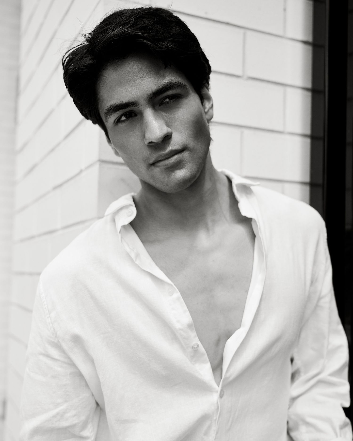 Black and white portrait of a young man with dark hair wearing an open white linen shirt, gazing confidently at camera.