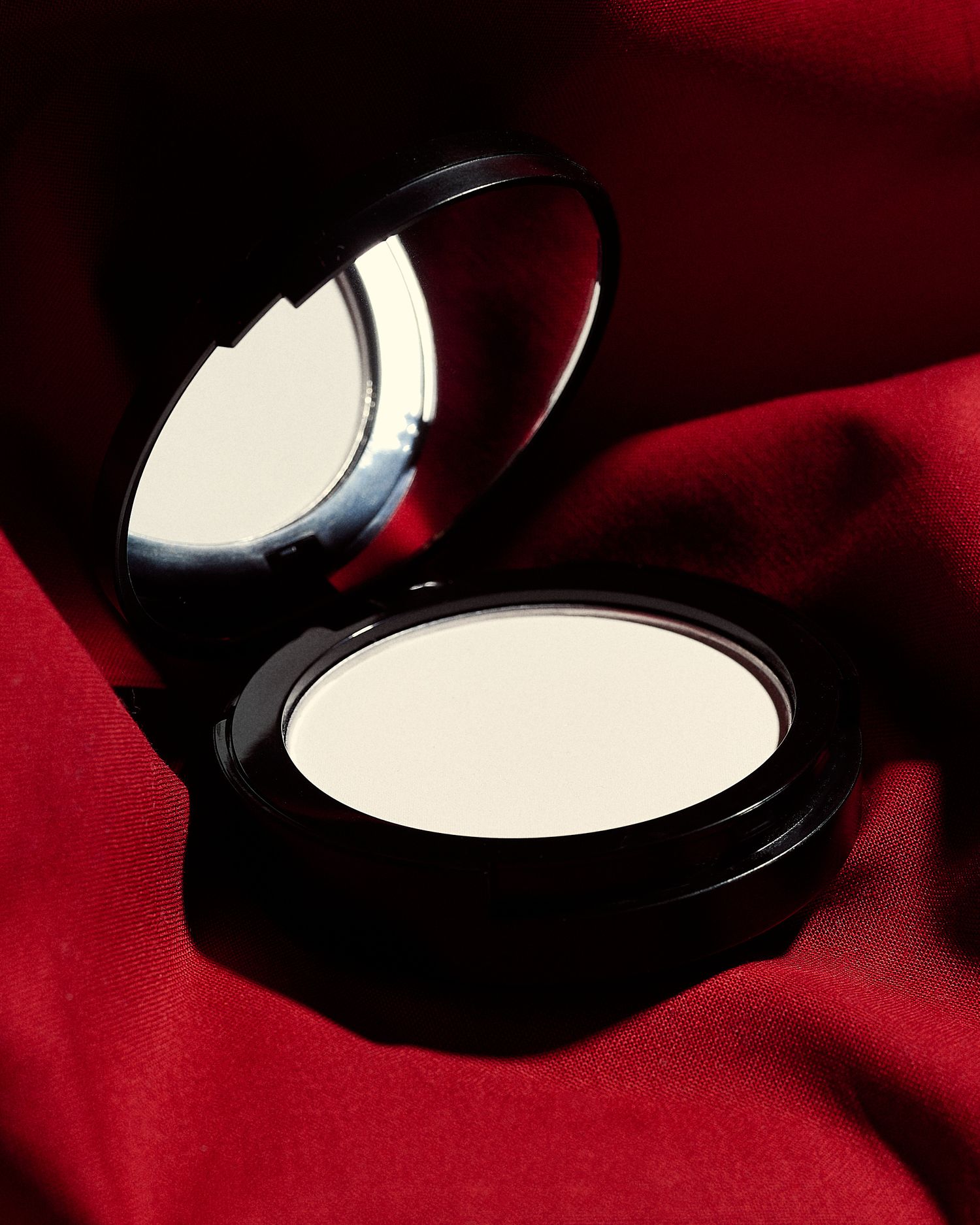 Open black compact powder case with mirror on red silk fabric background.