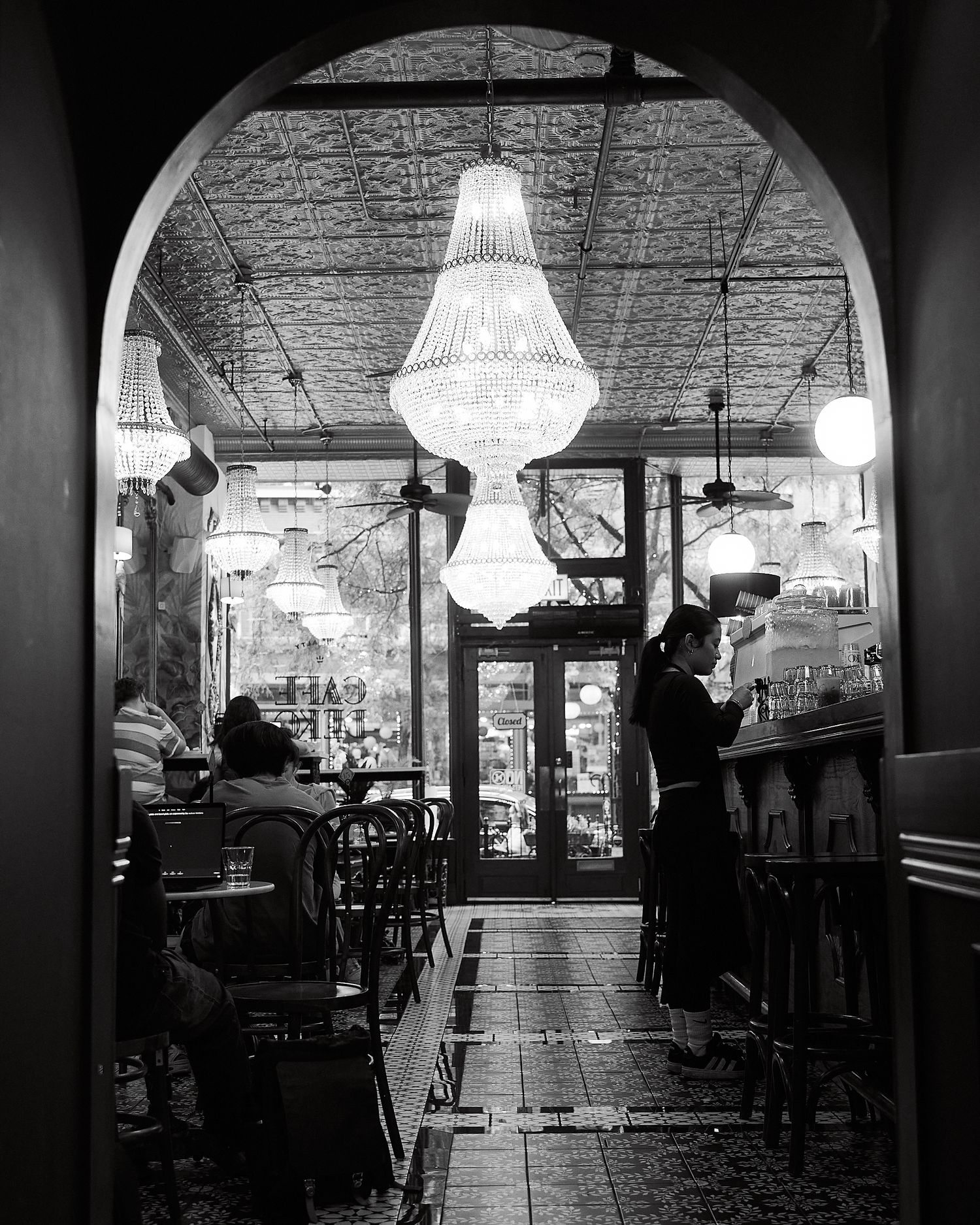 Black and white photo of elegant cafe interior with crystal chandeliers, arched doorway, and seated guests.