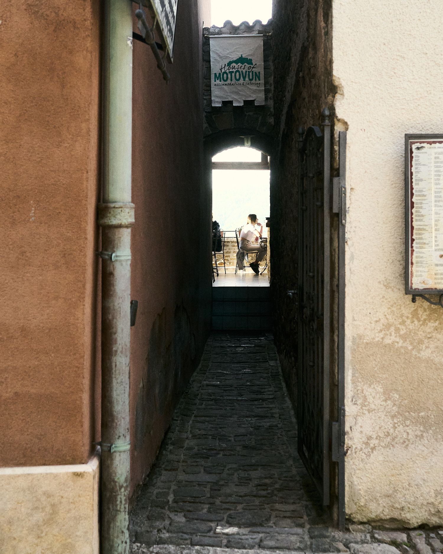 Narrow cobblestone alley between old stone buildings leading to an archway with a Motovon sign in a European town.