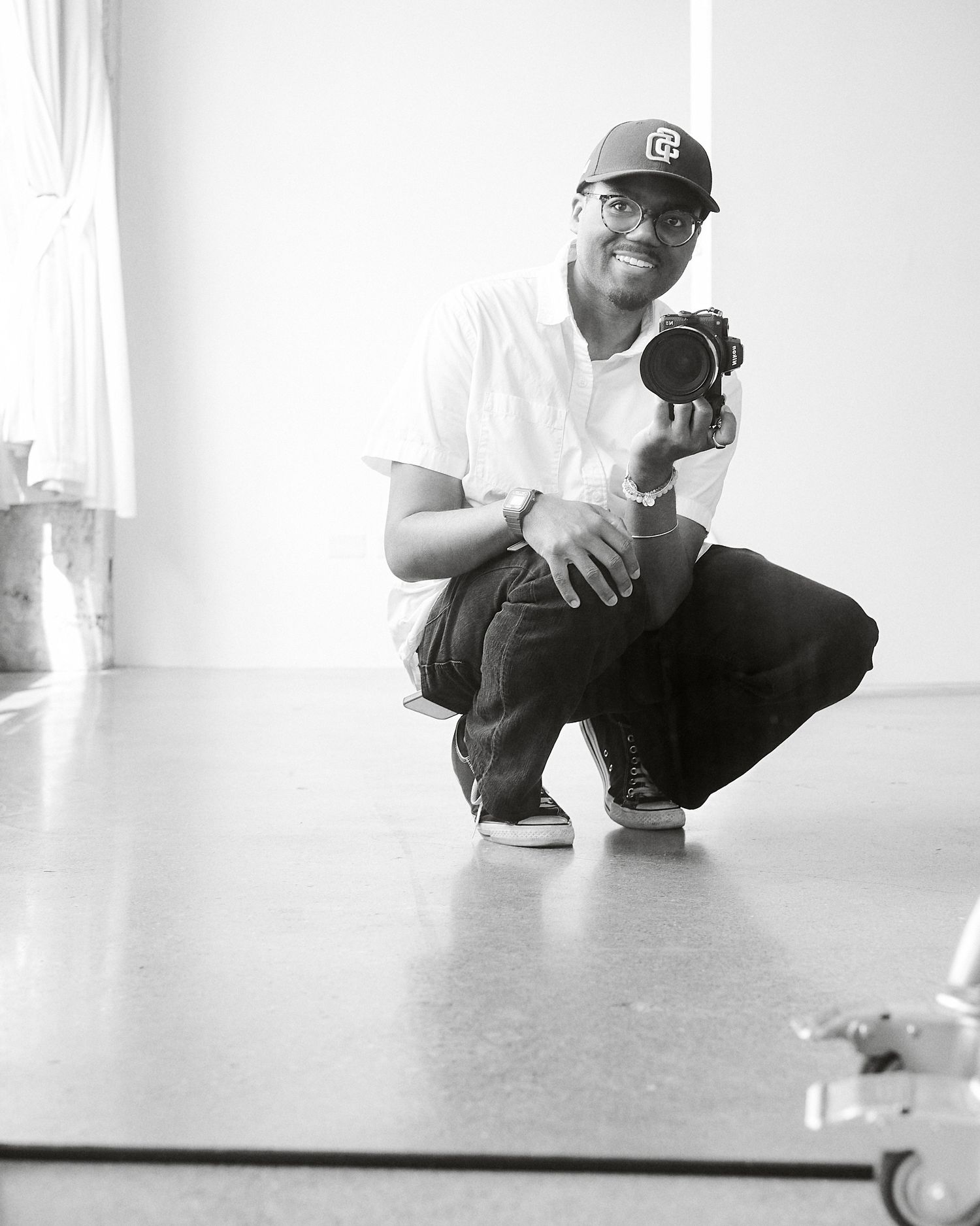 Black and white photo of a smiling man crouching while holding a camera in a bright studio setting.