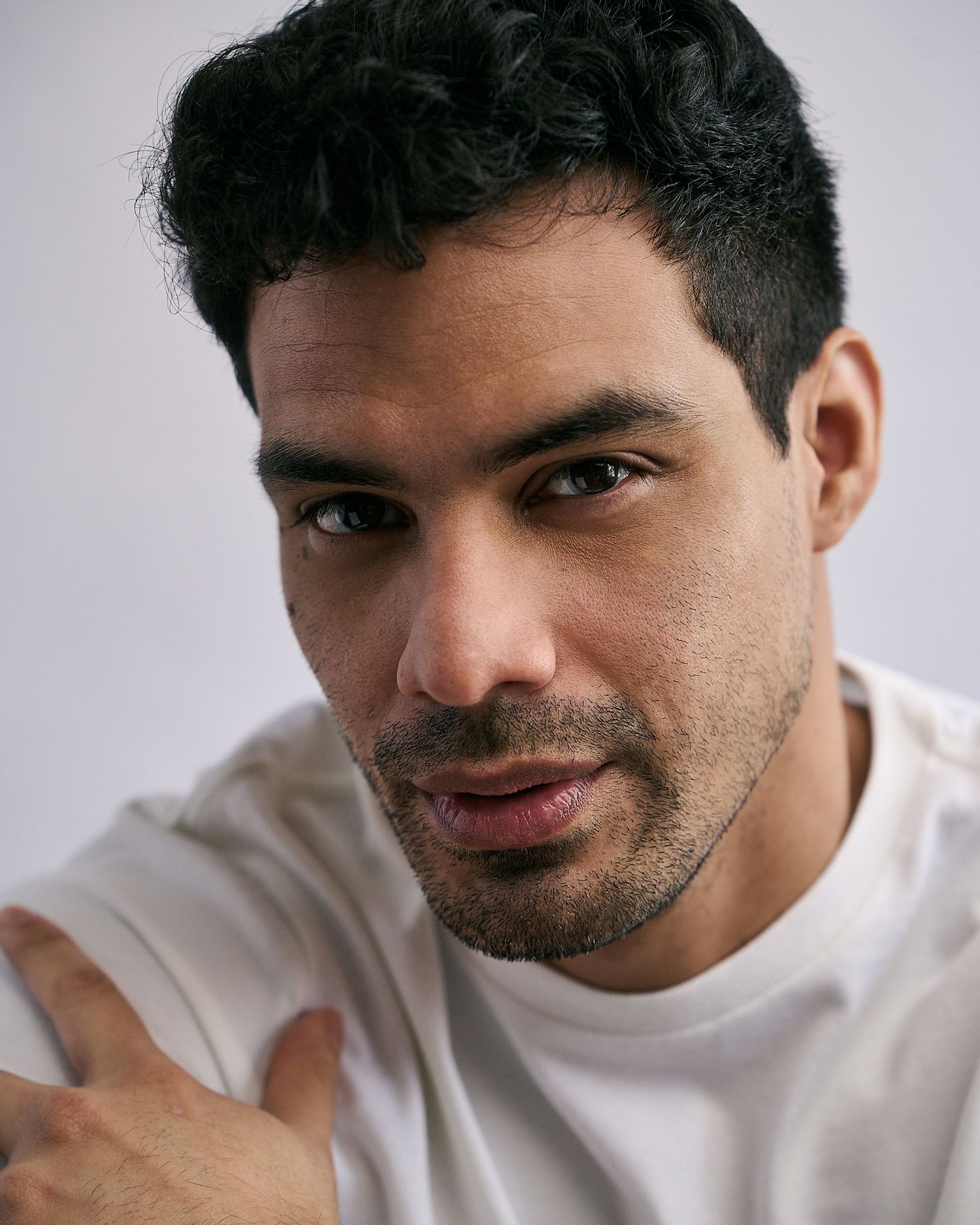 Close-up portrait of a young man with dark curly hair and light stubble wearing a white t-shirt.