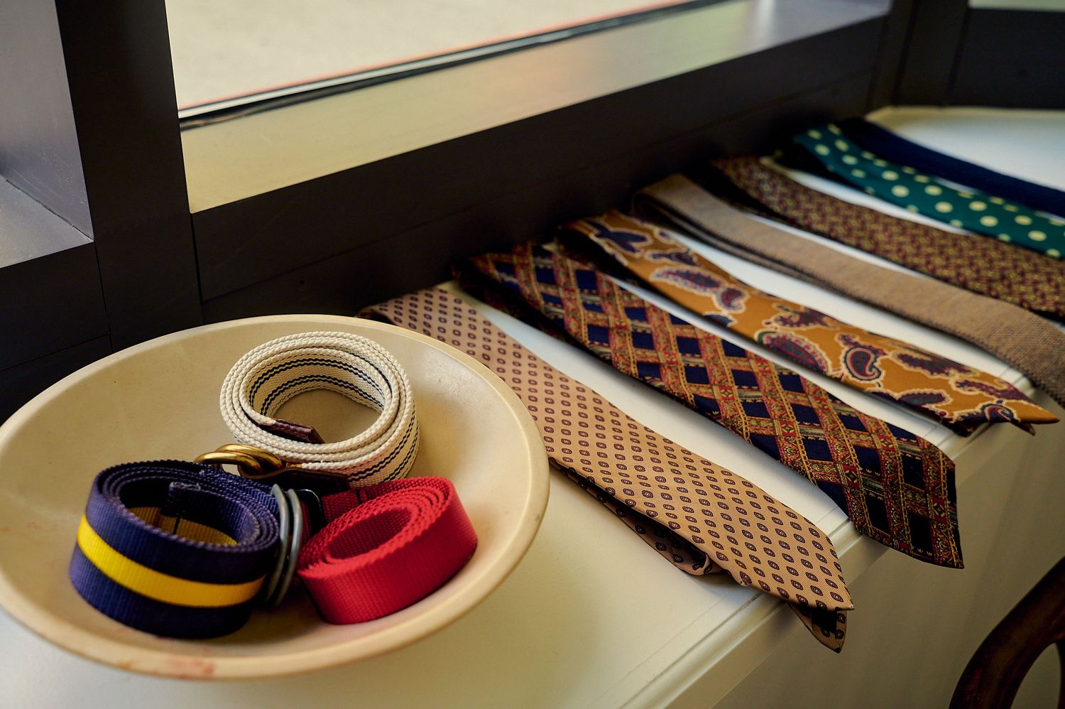 Colorful rolled belts in a bowl beside neatly arranged patterned neckties on a white drawer near a window.