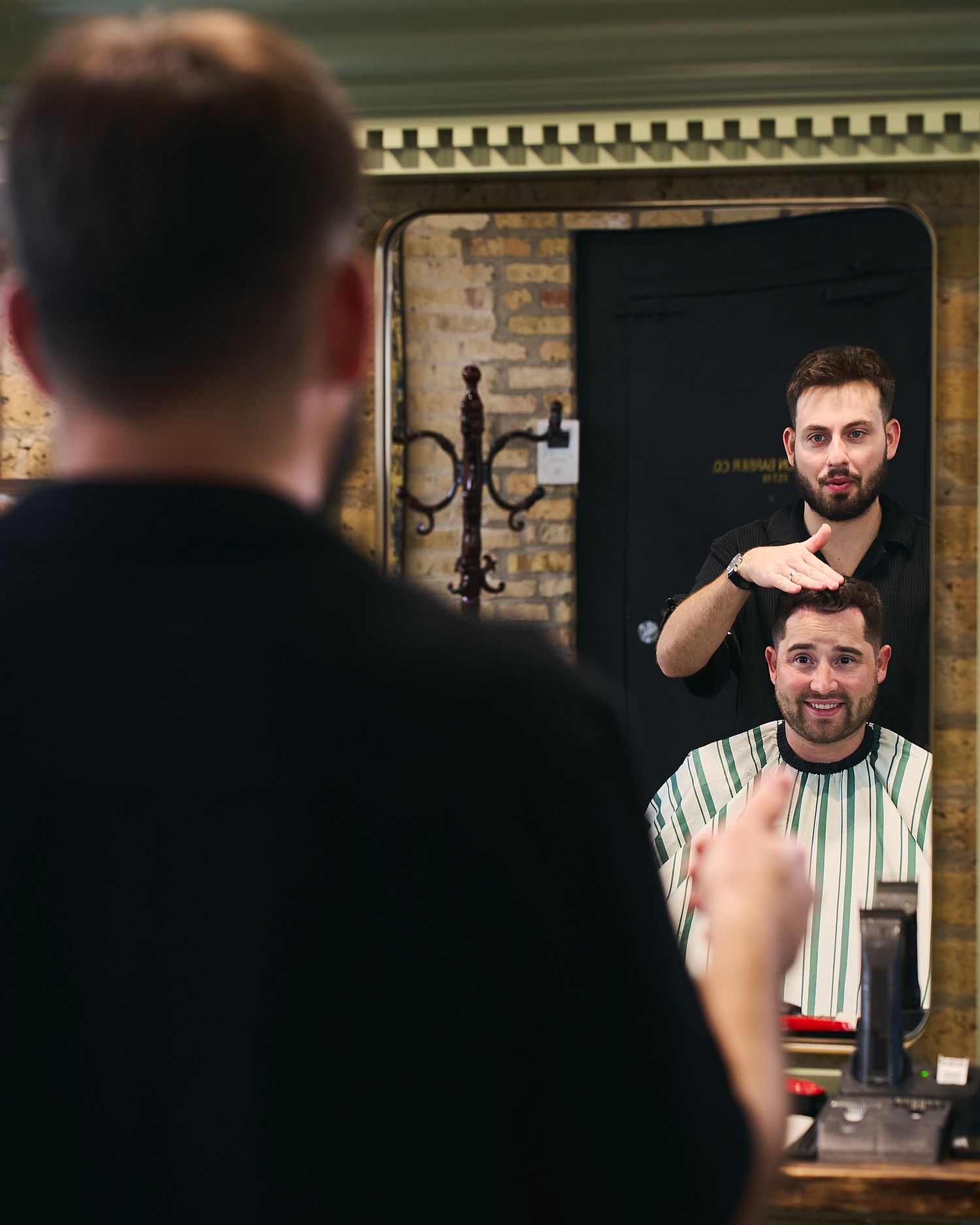Barber cutting clients hair reflected in a large ornate mirror in a traditional barbershop.