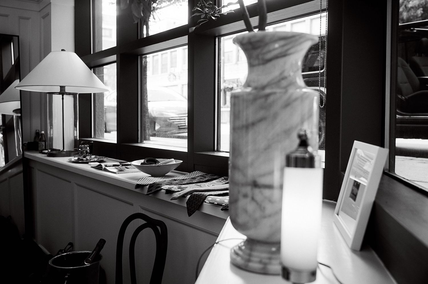 Black and white interior photo featuring a marble vase, lamp, and books on a desk near large windows.