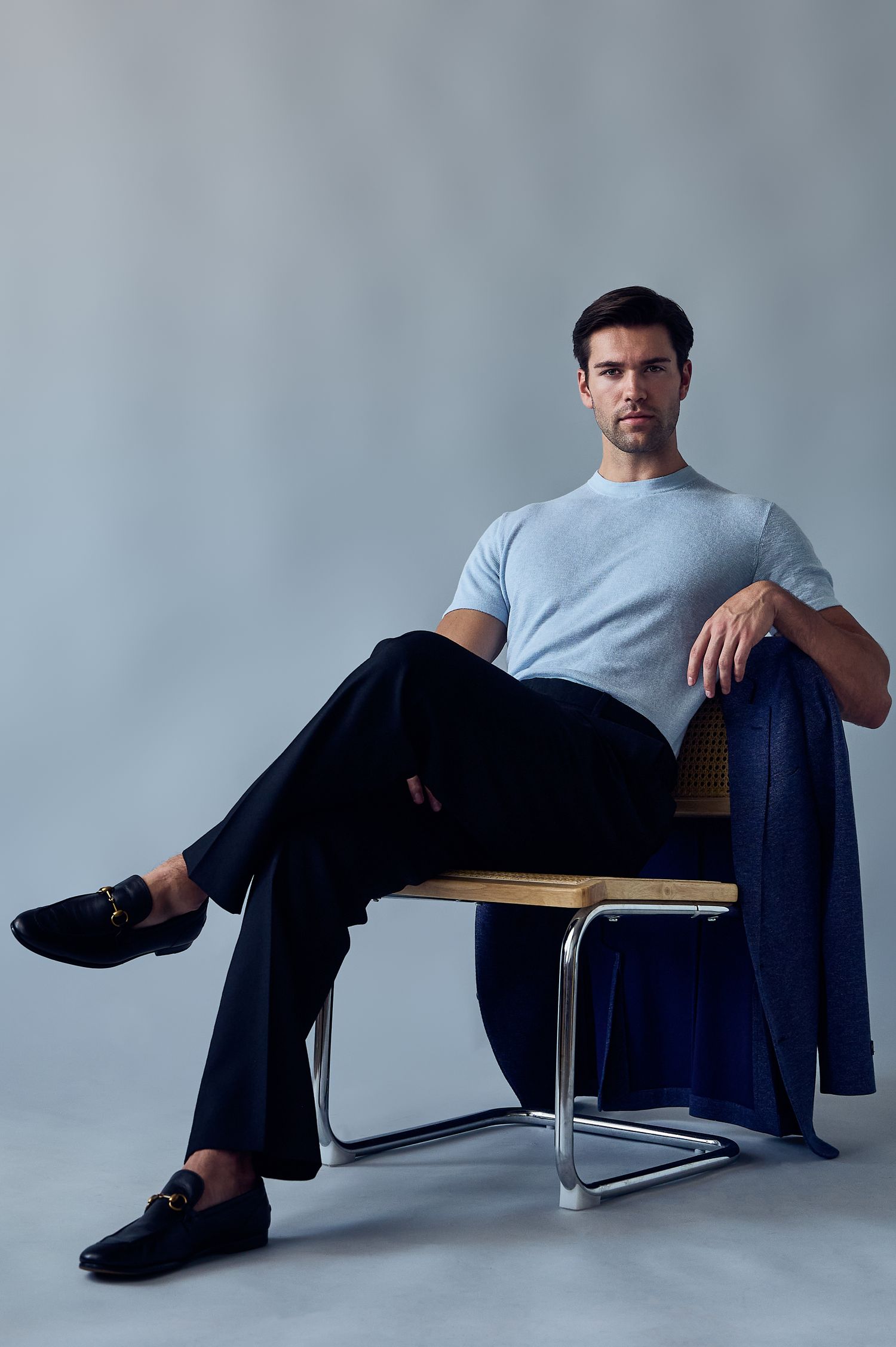 Man in light blue t-shirt and navy trousers seated on a modern chair with crossed legs and loafers.