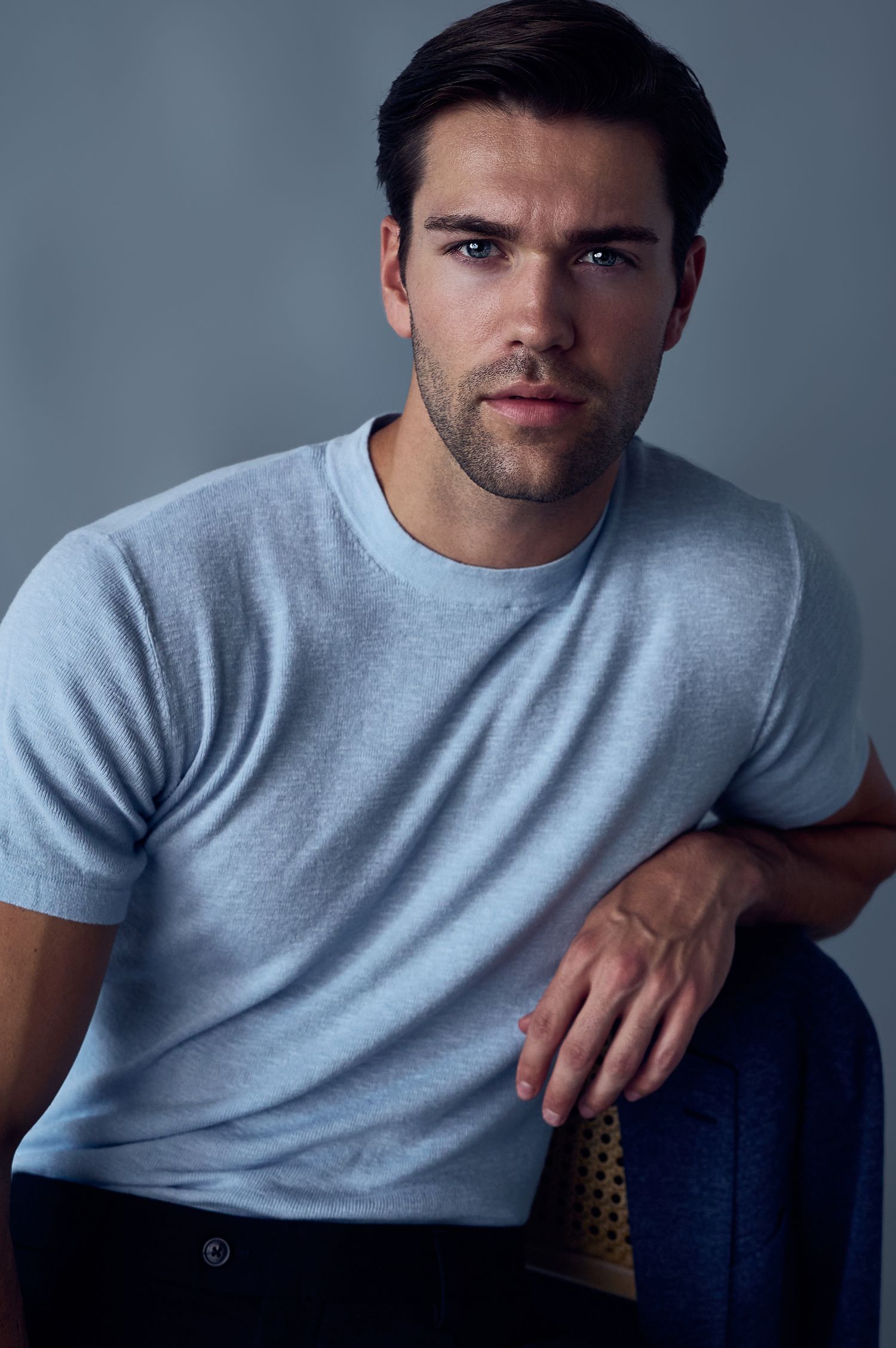 Young man with dark hair in a light blue t-shirt and jeans poses confidently in a studio setting.