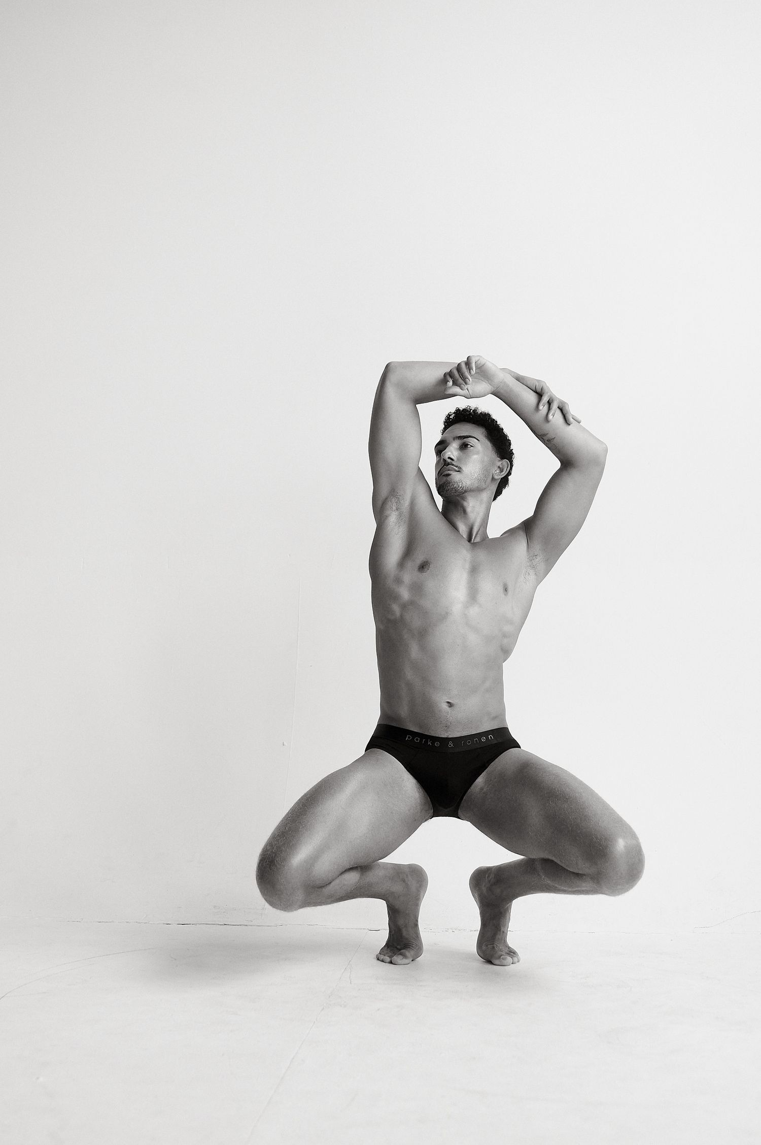 Muscular male model in black briefs performing a deep squat with arms raised overhead in a white studio.