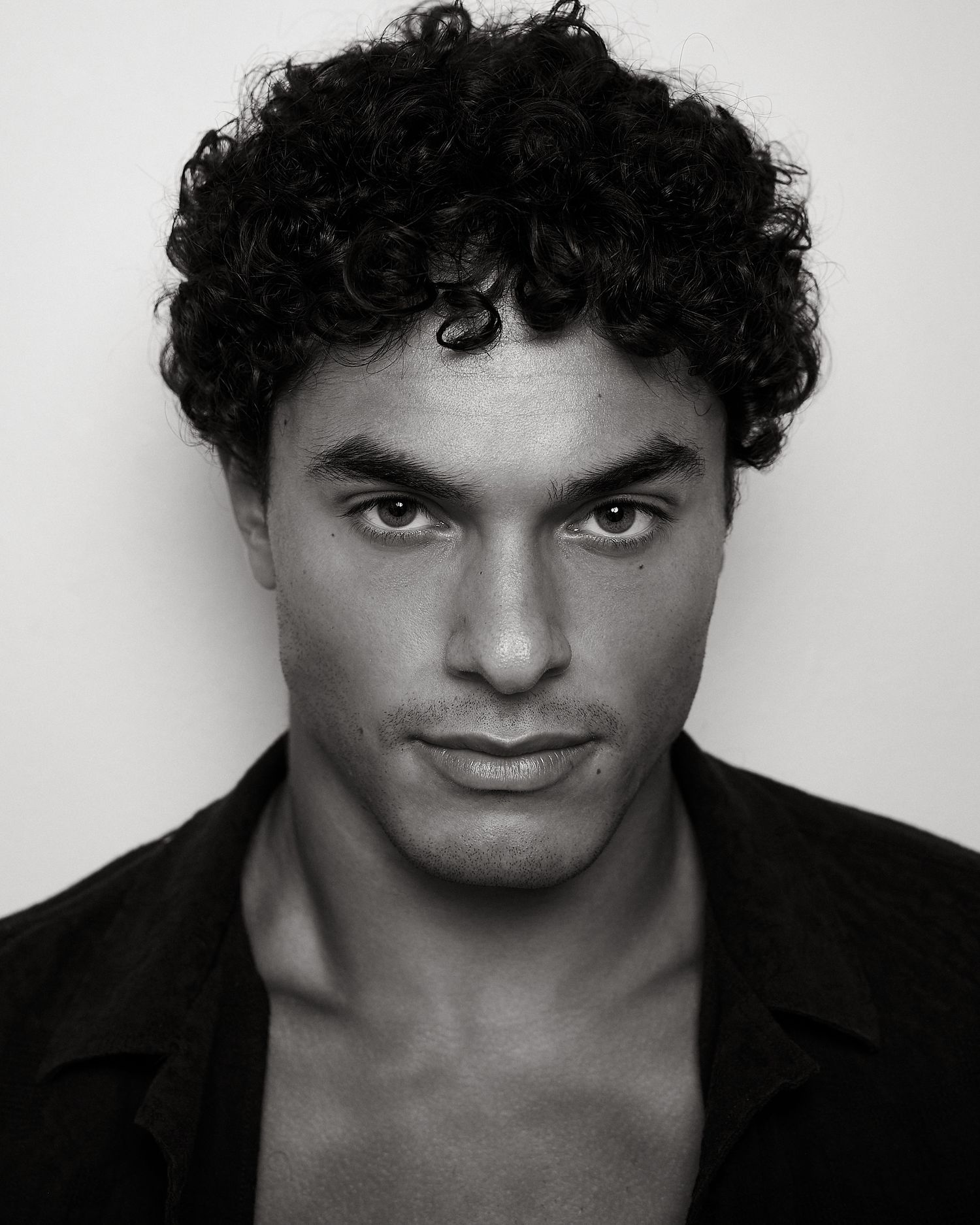 Black and white close-up portrait of a man with curly hair and an open dark shirt, intense gaze.