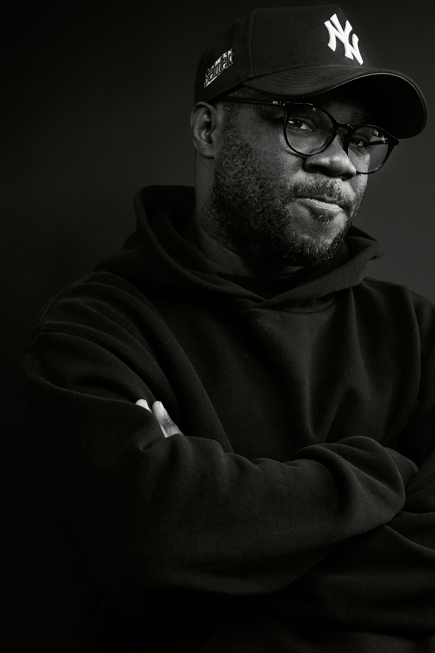 Serious man in NY Yankees cap, glasses and black hoodie with arms crossed in dramatic low-key portrait.