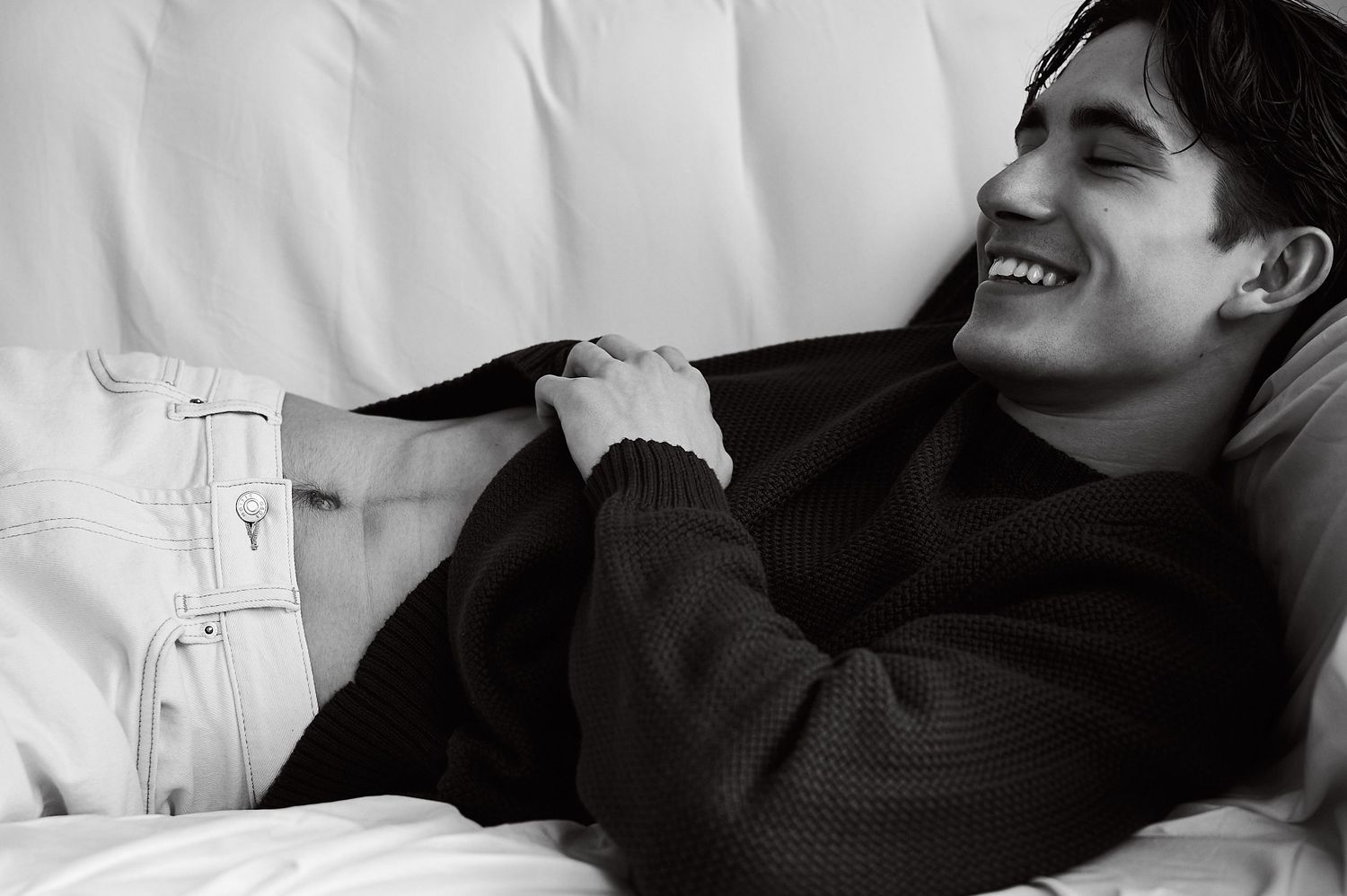 Young man laughing joyfully while relaxing on a white sofa, wearing a dark knit sweater, black and white photo.