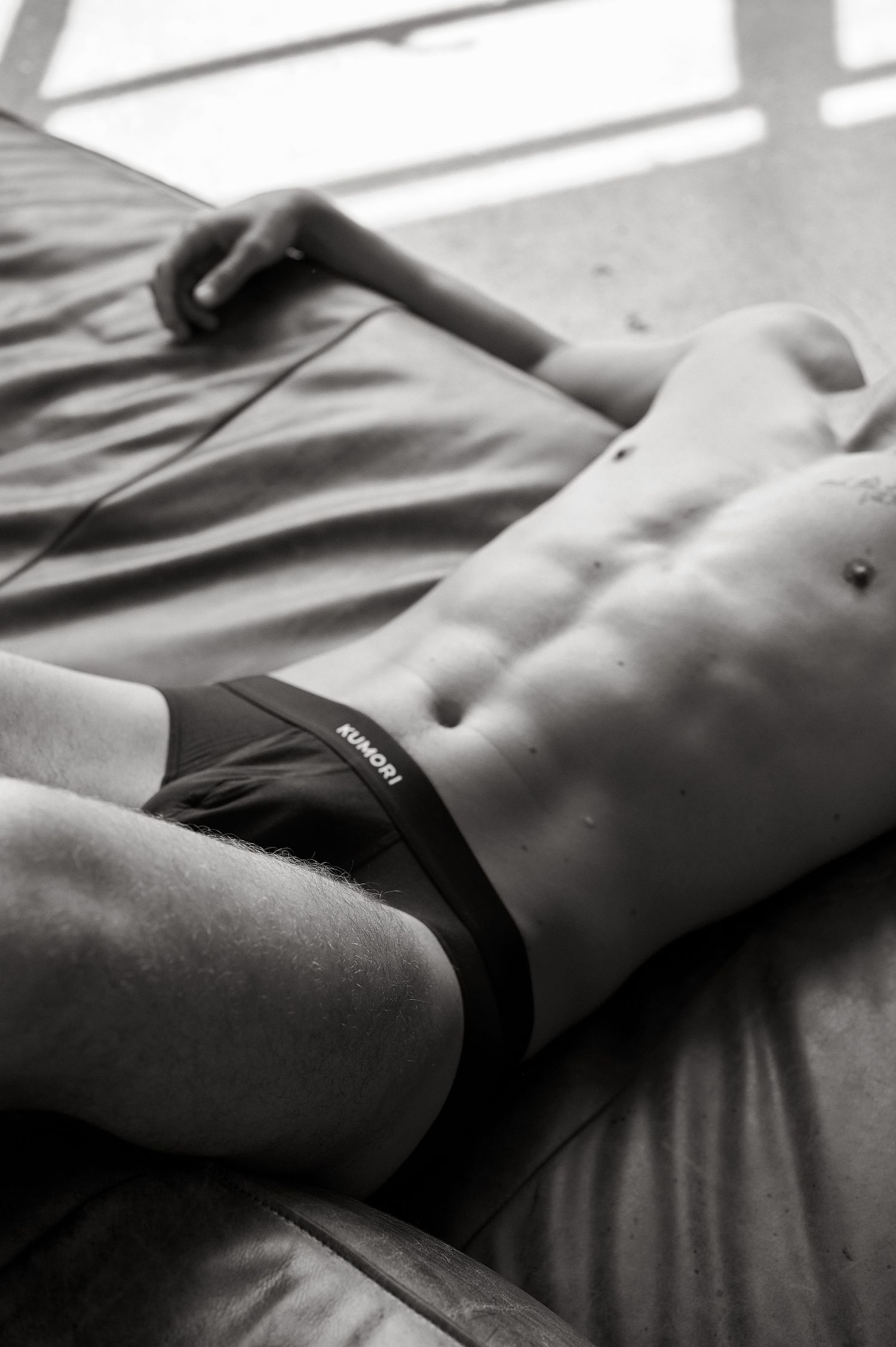 Black and white photo of a fit male torso wearing Kimchi black briefs, lying on a bed.