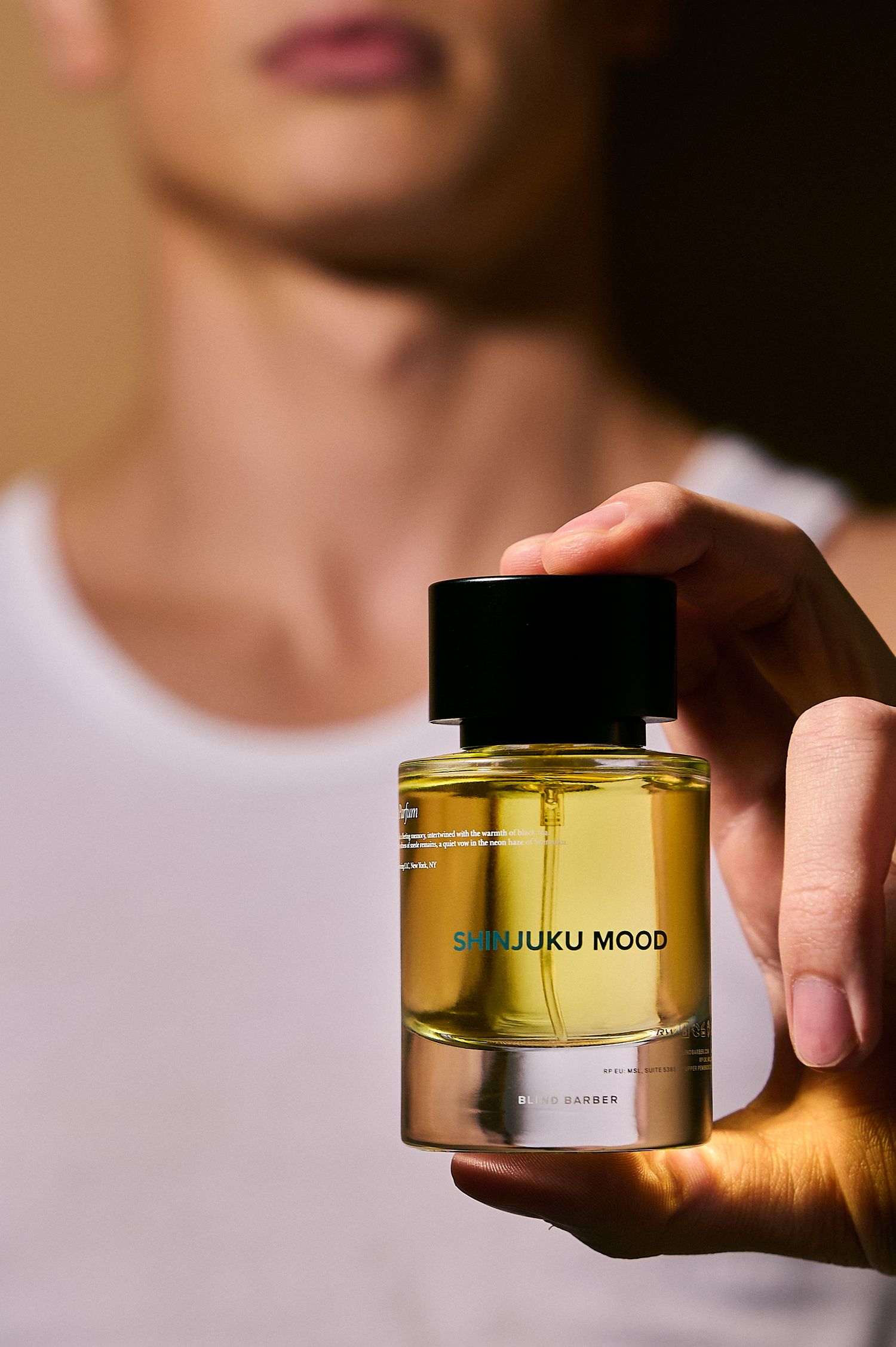 Hand holding Shinjuku Mood fragrance bottle in foreground with blurred figure in white tank top.