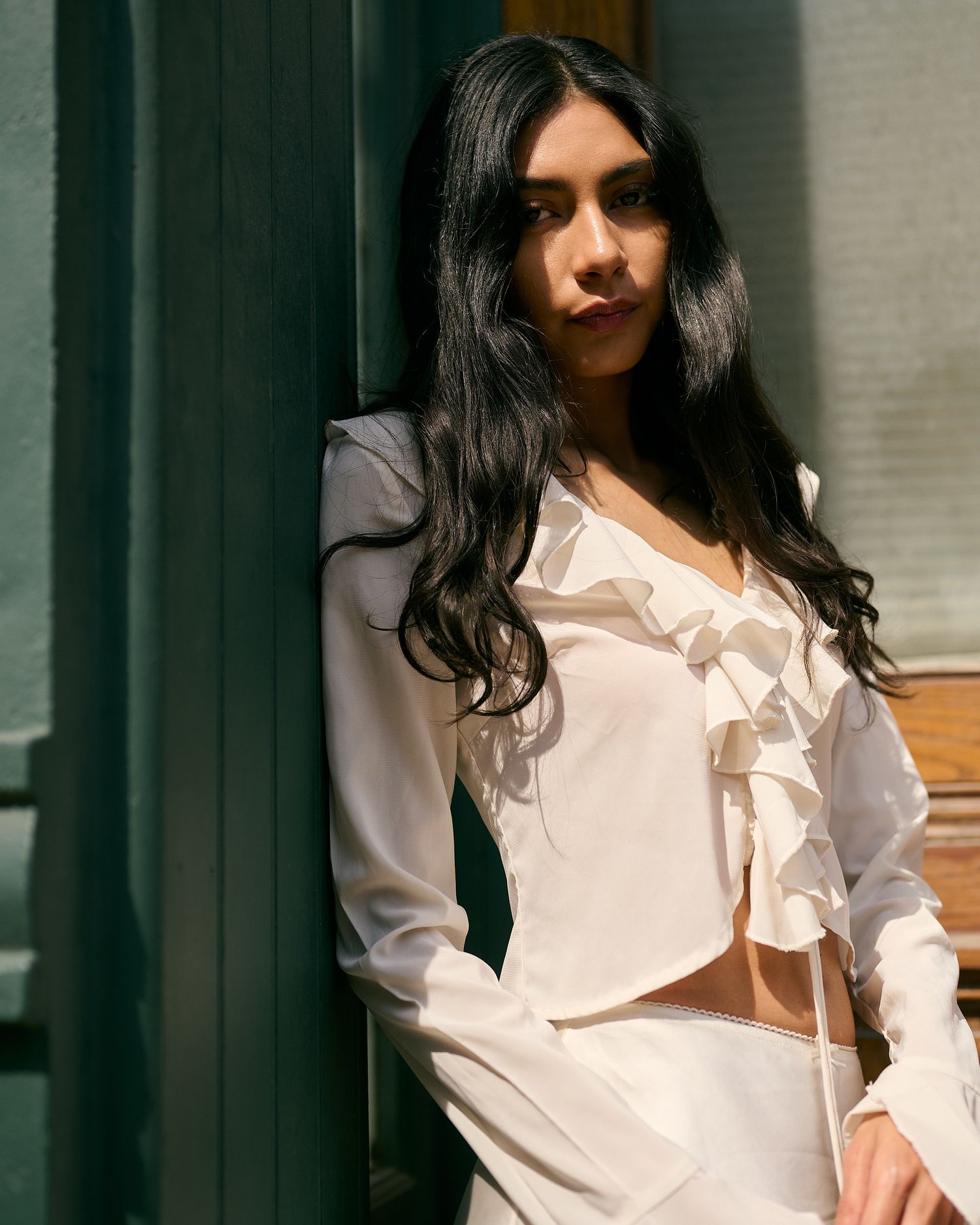 Woman with long dark hair wearing a white ruffled sheer blouse leaning against a green doorframe.