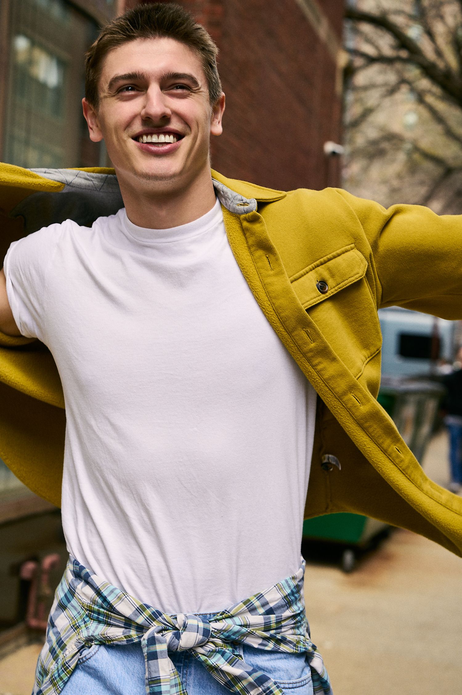 Smiling young man in white tee, mustard jacket, and plaid shirt tied at waist, posing outdoors on urban street.