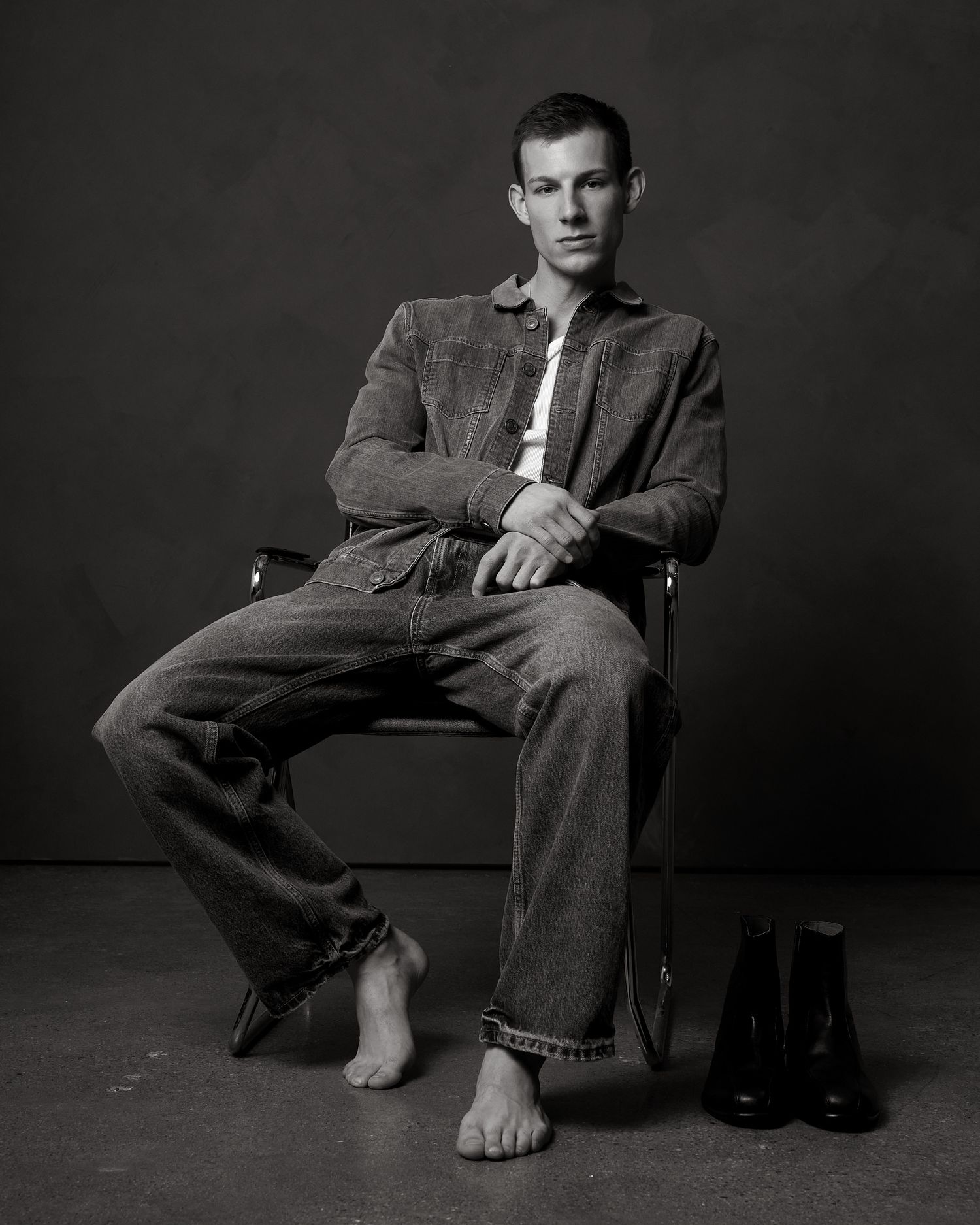 Black and white portrait of a barefoot man in denim jacket and jeans seated in a chair with boots beside him.