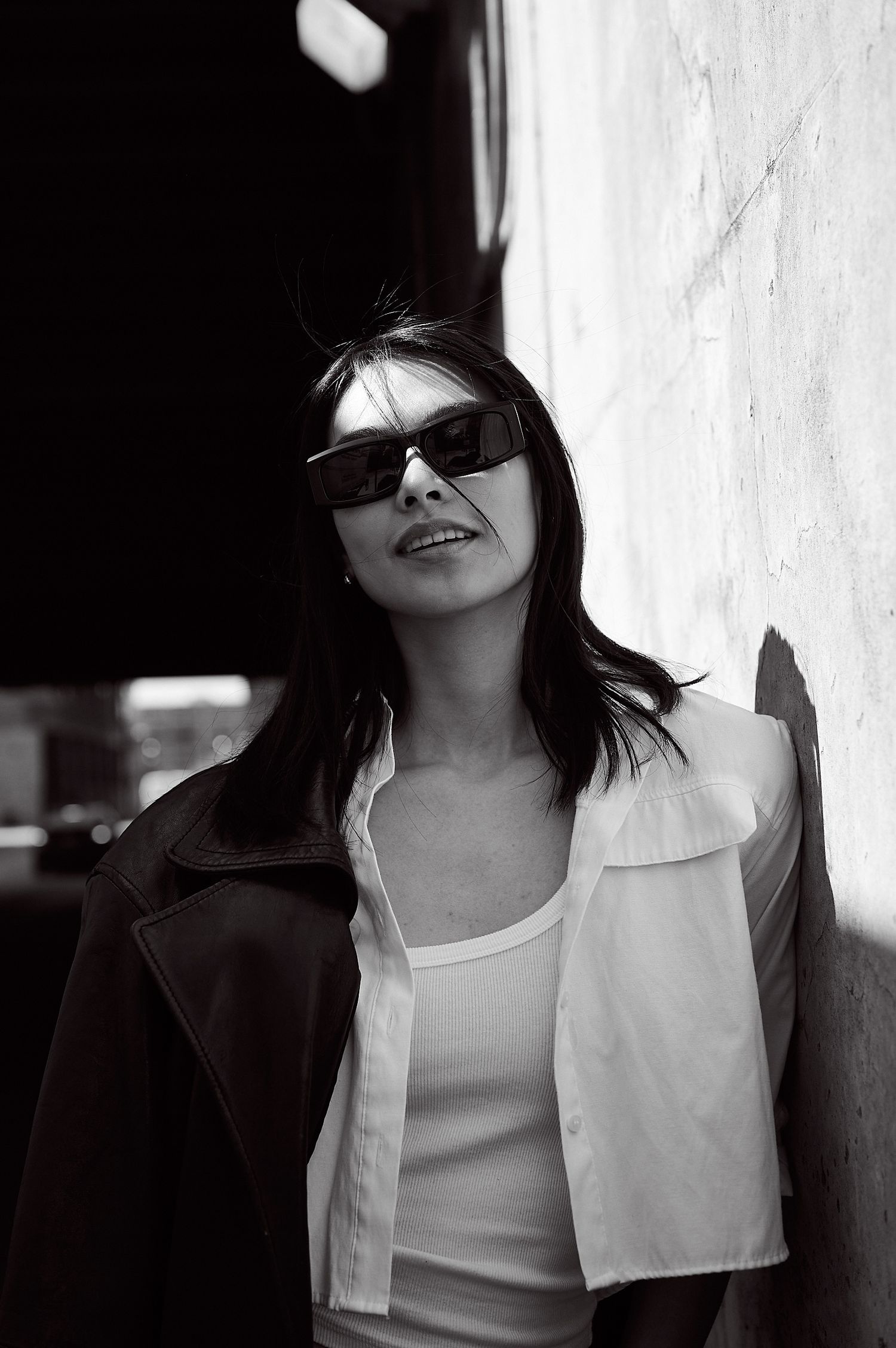 Black and white portrait of a woman in sunglasses, white shirt, and dark jacket leaning against a wall.