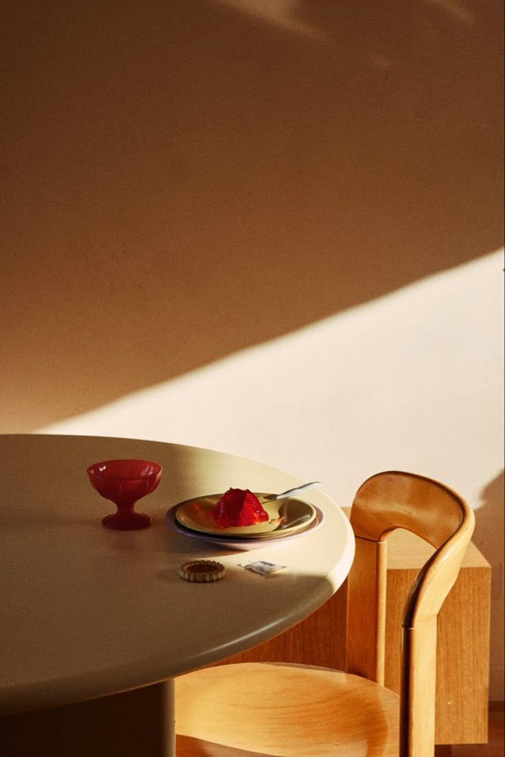 Minimalist dining table setting with red dessert, decorative bowl, and wooden chair bathed in warm afternoon sunlight.