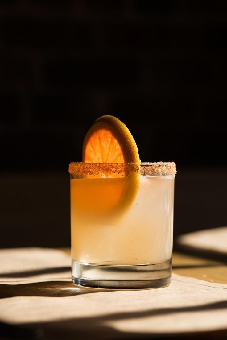 A spiced-rim cocktail glass with orange slice garnish, golden drink illuminated by dramatic sunlight on wooden surface.