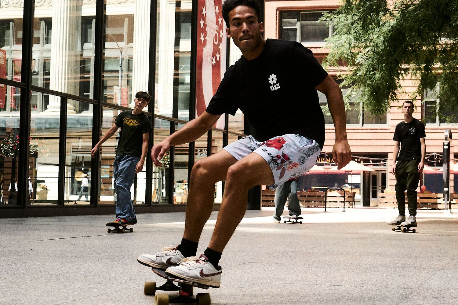 Young man skateboarding on a longboard in an urban plaza wearing floral shorts and black tee.