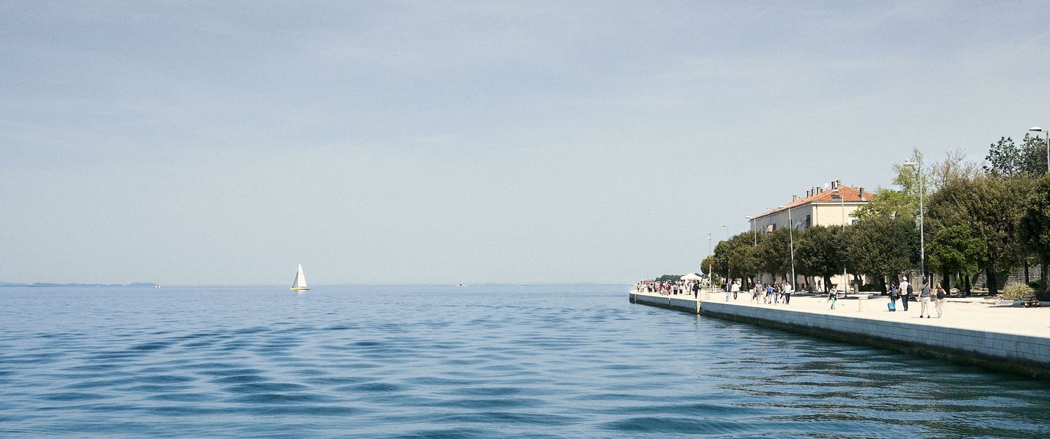 Calm blue sea with a sailboat on the horizon beside a tree-lined coastal promenade on a clear day.