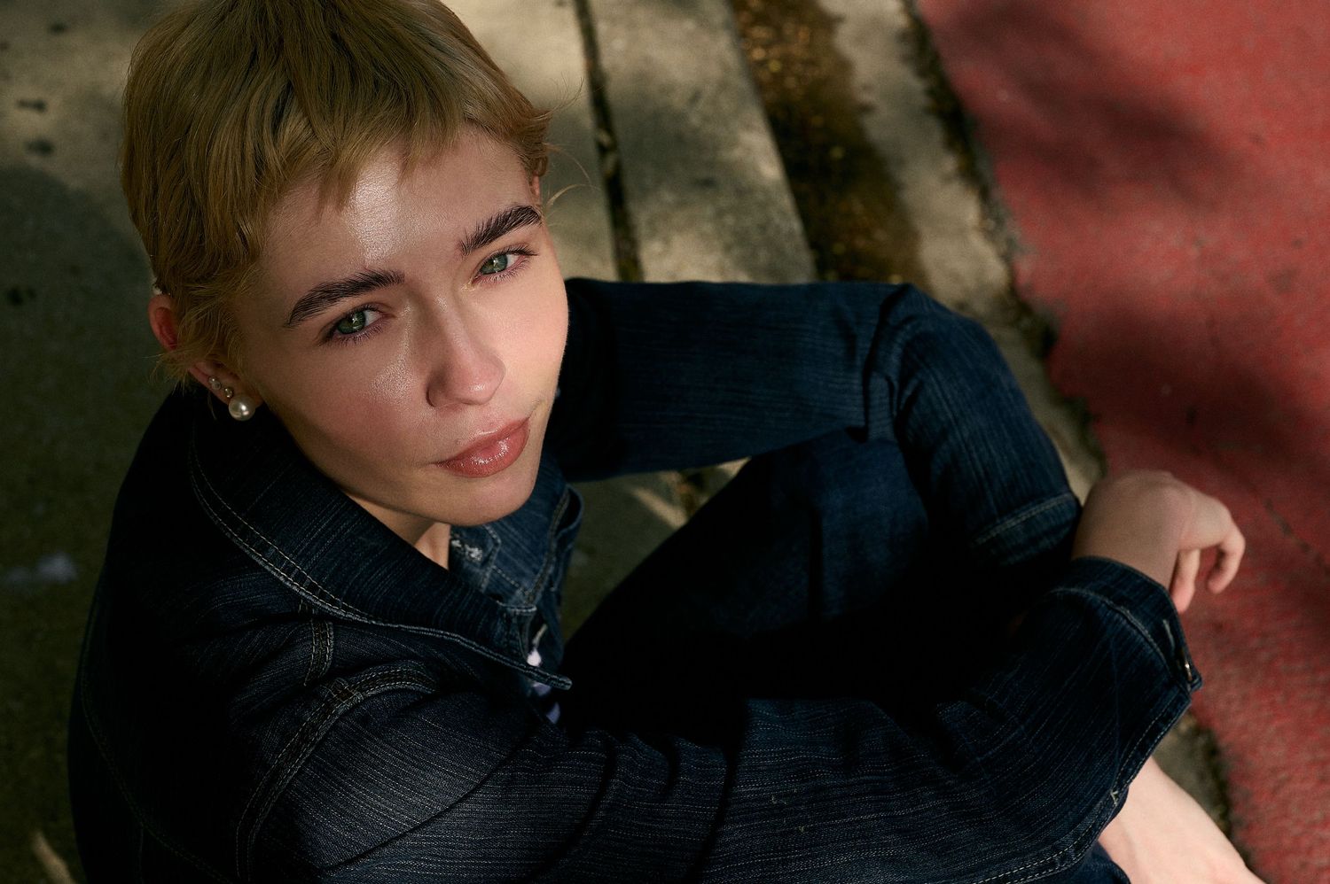 Young person with short blonde hair wearing a denim jacket sitting on the ground looking up at the camera.