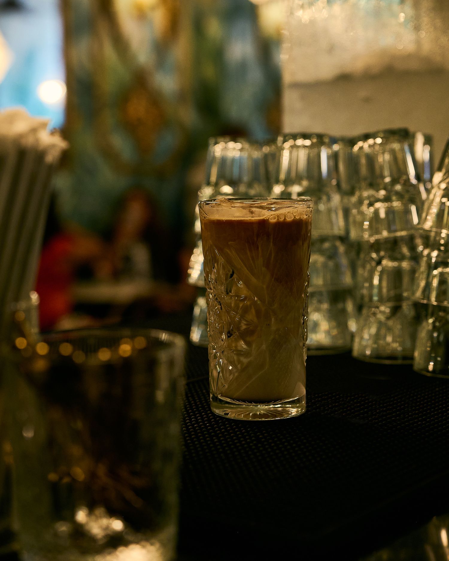 A decorative glass filled with an iced coffee drink sits on a dark surface surrounded by empty glasses in a dimly lit bar.