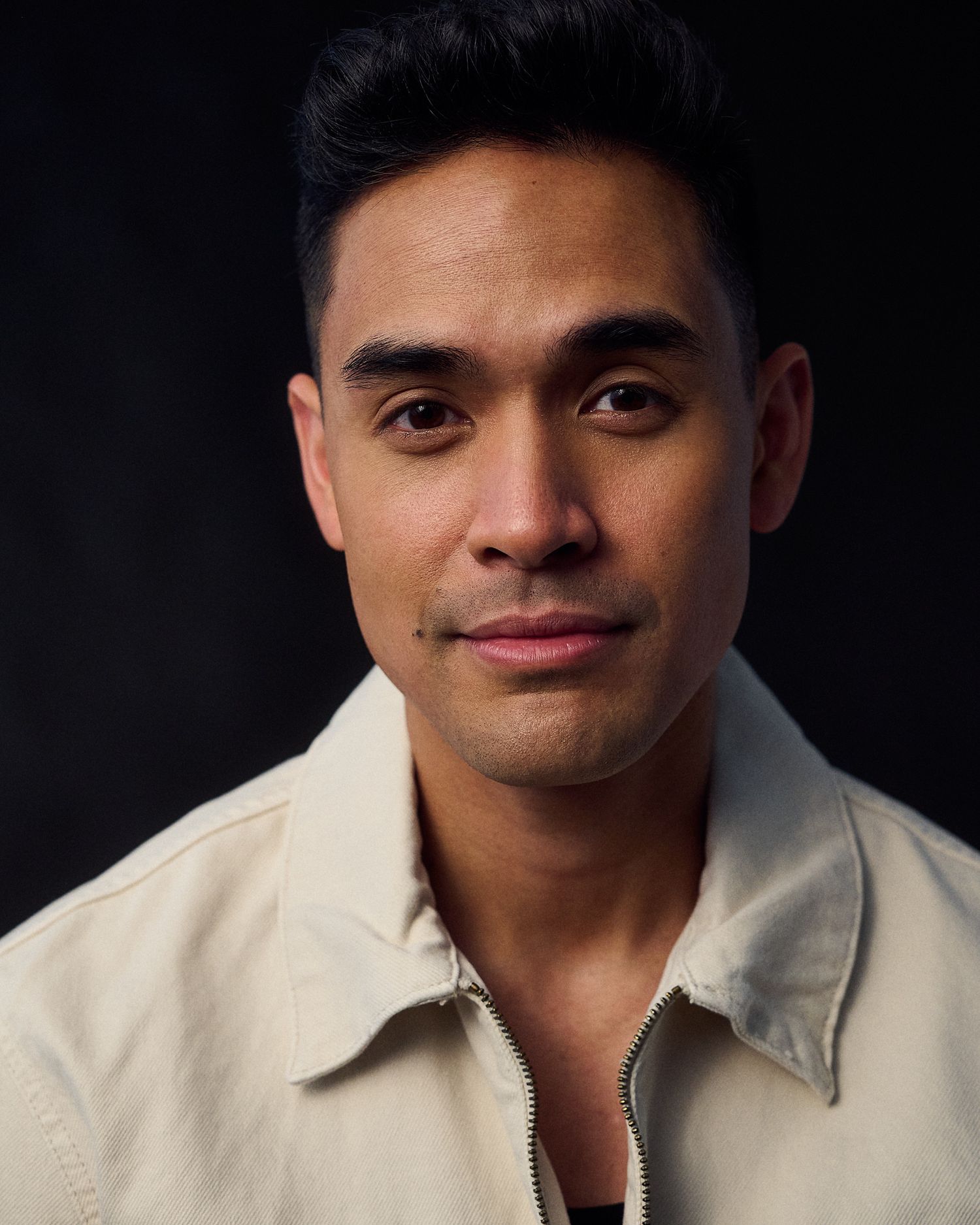 Professional headshot of a young man in a cream zip-up jacket against a dark background.