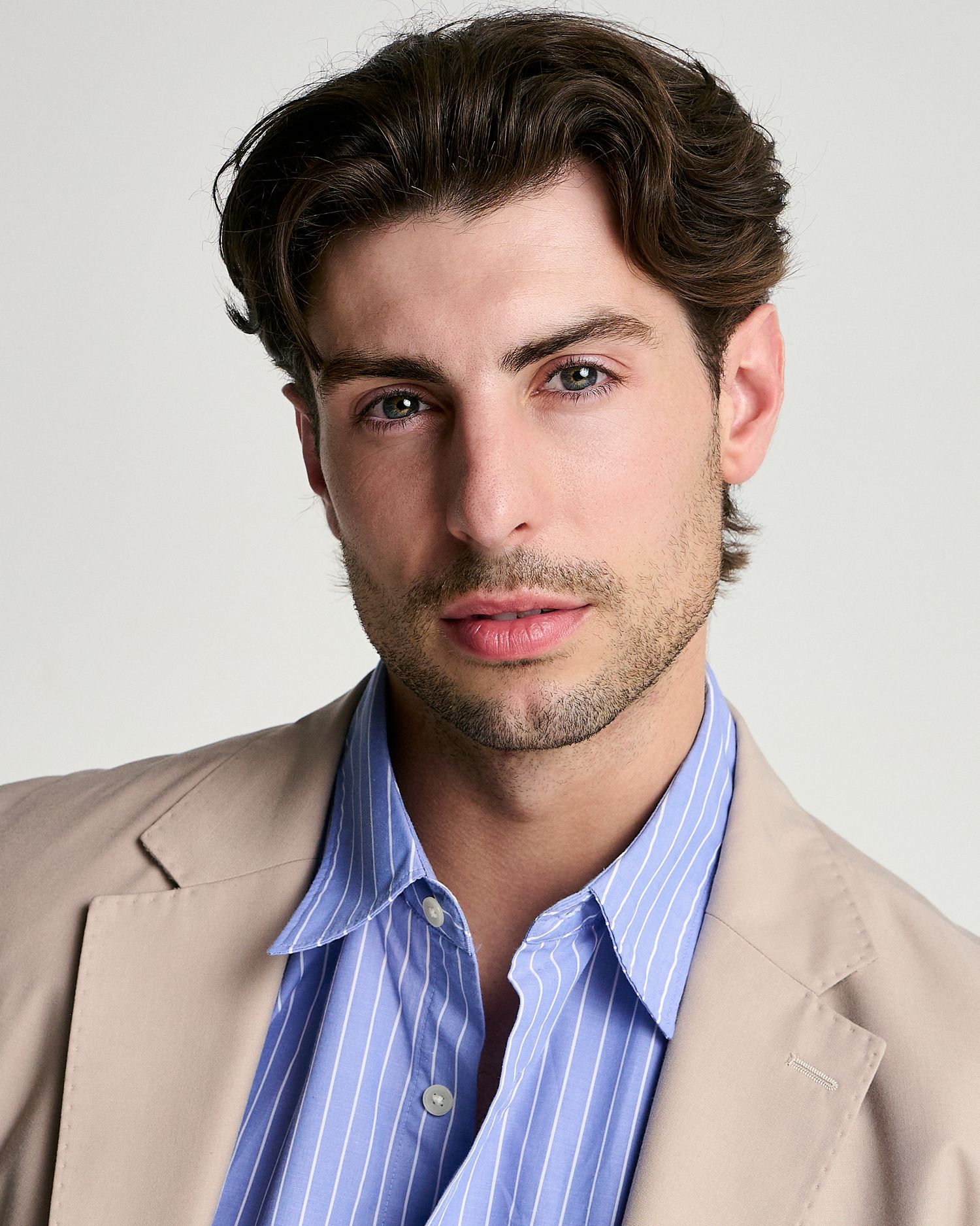 Young man with dark wavy hair wearing a blue striped shirt and beige blazer poses for a professional headshot.