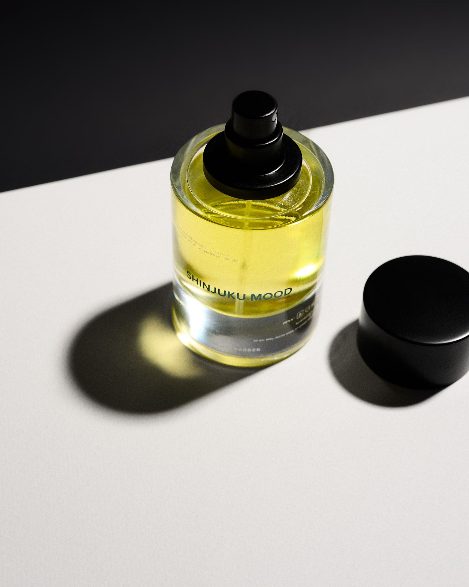Yellow Shinjuku Mood perfume bottle with black cap on white surface against dark background.