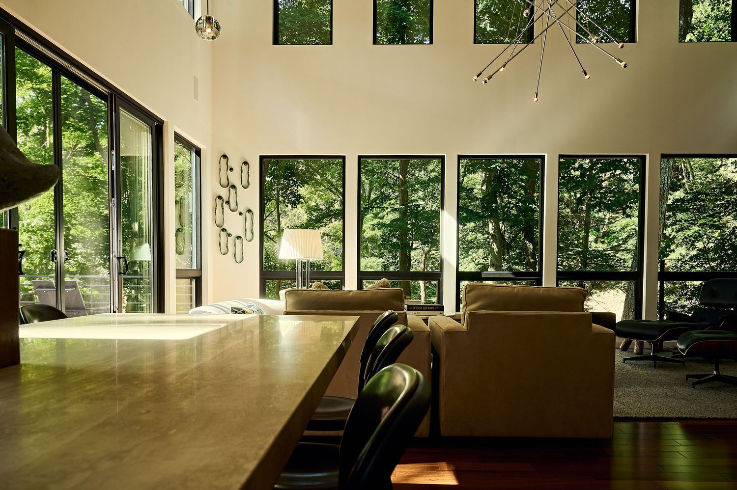 Modern open-plan living room with floor-to-ceiling windows overlooking lush green trees, featuring leather sofas and wooden surfaces.
