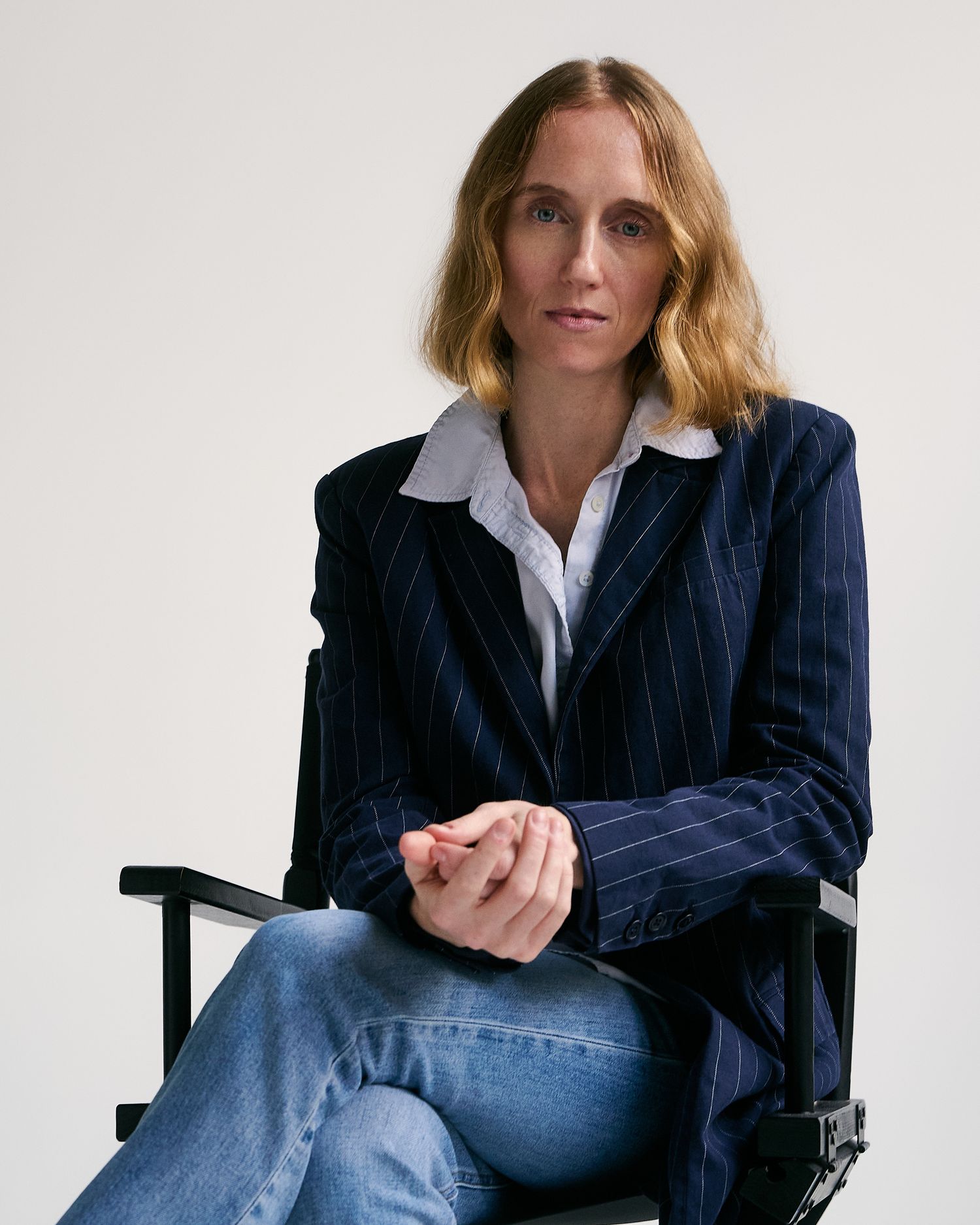 Woman with blonde hair wearing a navy pinstripe blazer seated in a director's chair, hands clasped.